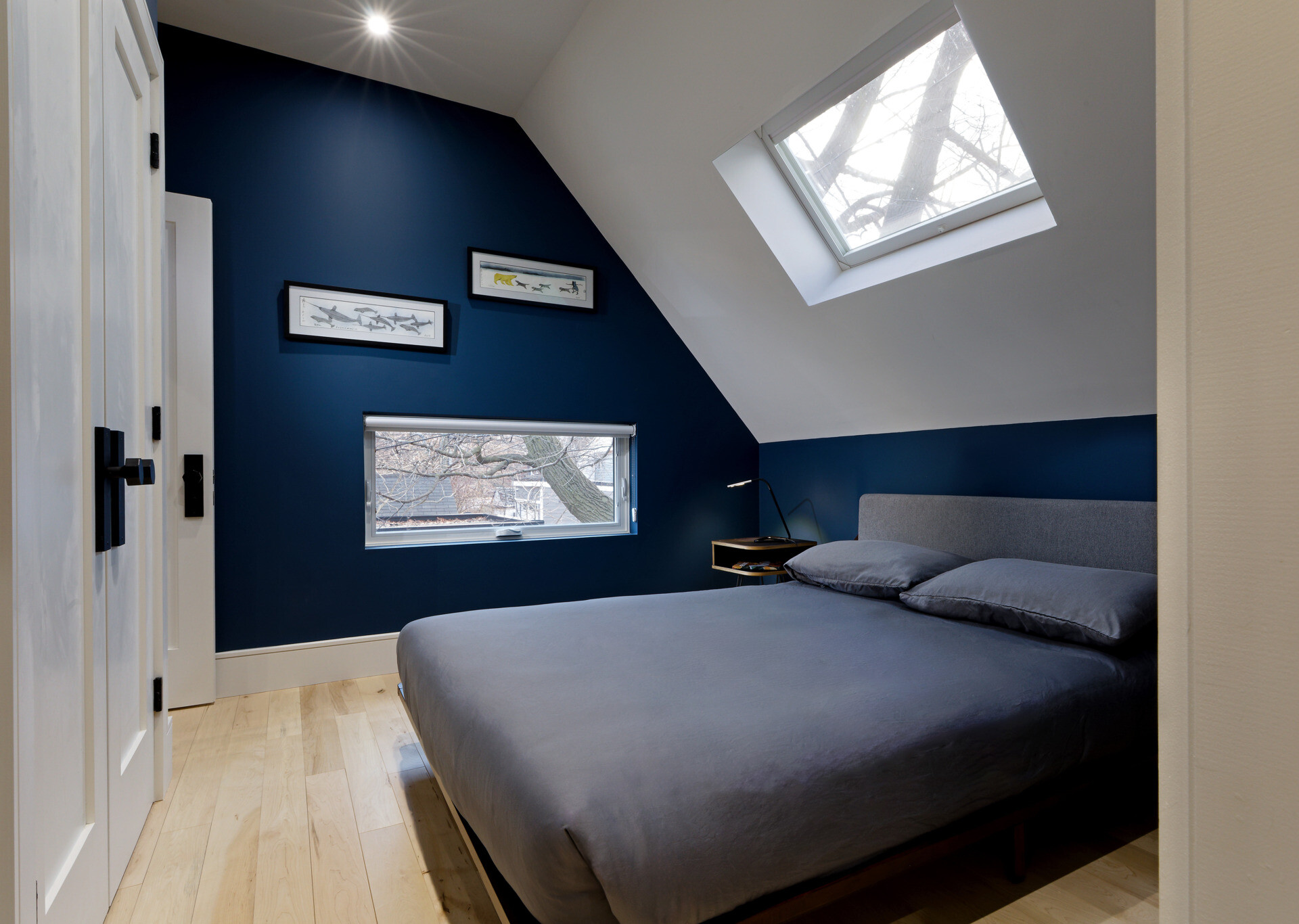 In this bedroom, walls in deep blue contrast with a white sloped ceiling, creating a serene cocoon under the canopy of the house. A horizontal window and a skylight above the bed bring the sky into view, while a built-in closet maximizes storage.