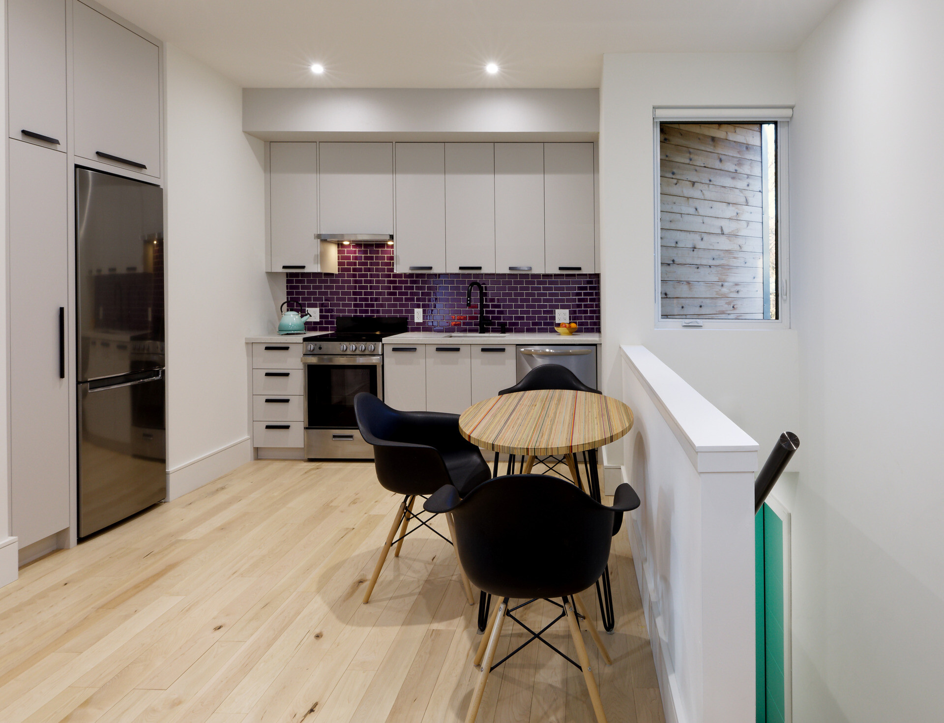 The dining area of this modern laneway house features a round wooden table with two chairs, framed by soft light that spills in from multiple angles. Just beyond, the kitchen balances subtlety with surprise: light grey minimalist cabinetry, matte black hardware, and a backsplash of glazed purple subway tiles that shimmer like amethyst.