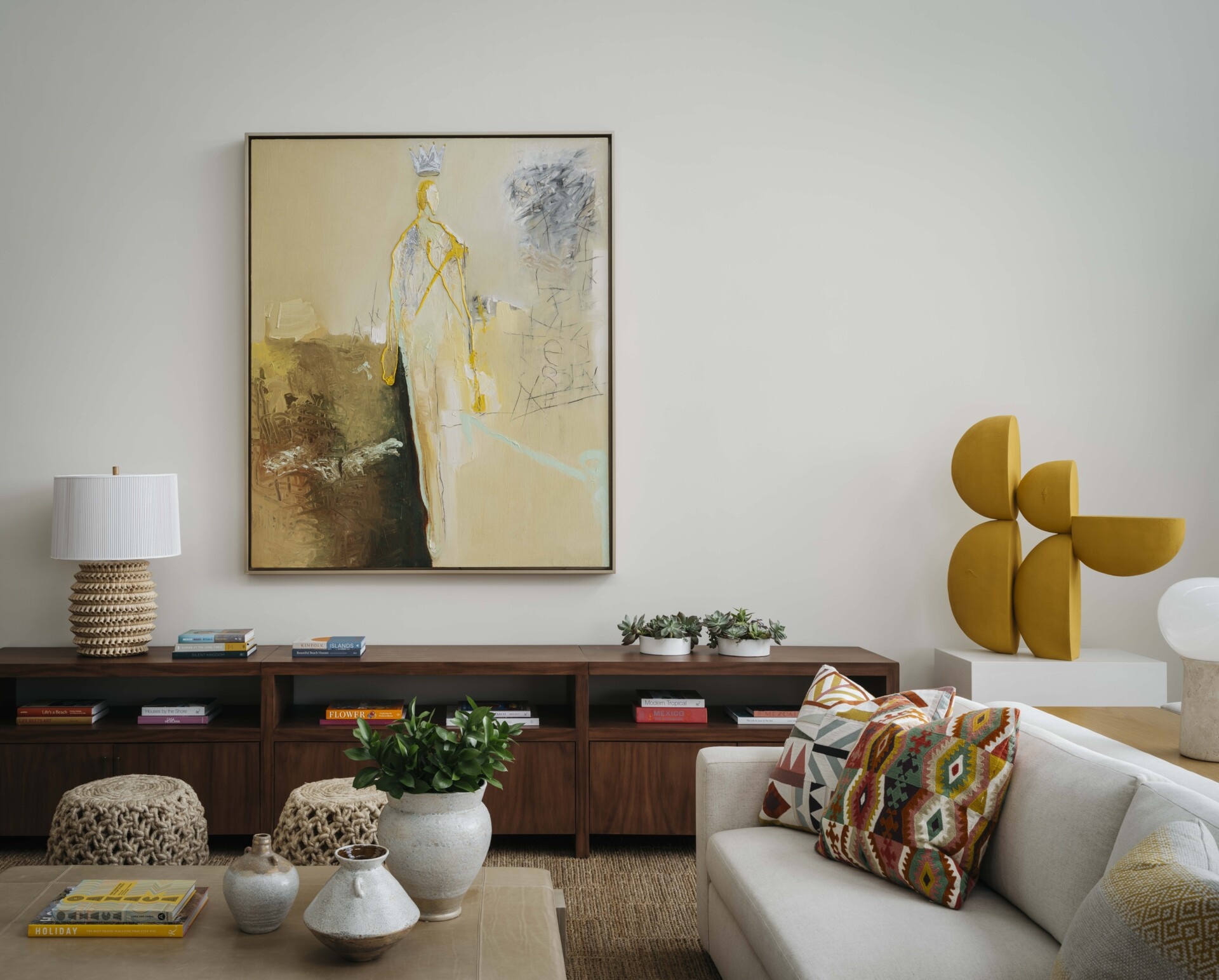 The living room is a study in contrast, its neutral, modern shell becomes a gallery for rich textiles, collected art, and sculptural lighting. Anchored by a bold Frank Arnold painting sourced locally in San Jose del Cabo, the space welcomes conversation and lounging in equal measure. Natural textures like seagrass and rope weave through ottomans and rugs, grounding the room in regional craft traditions. Soft, neutral-toned upholstery allows the vivid accents to take center stage, making the living space feel layered yet light.