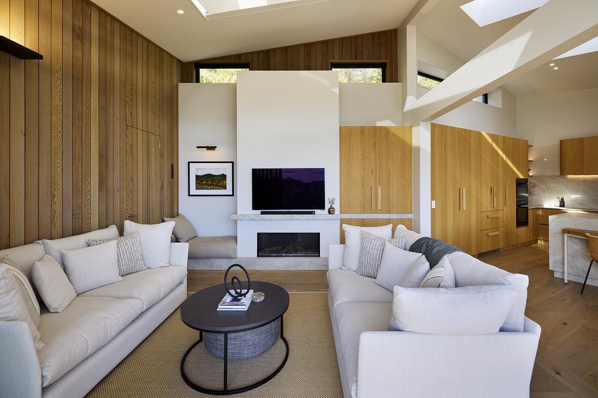 This living room combines natural textures with a clean, open layout. Vertical timber paneling brings warmth and contrast to the white walls and vaulted ceiling. Stone-toned sofas offer comfortable seating, anchored by a dark oval coffee table. The fireplace wall includes a built-in TV, with a bench and art niche tucked to the side for reading or relaxing. Skylights and clerestory windows fill the room with light, keeping the space bright and uncluttered.
