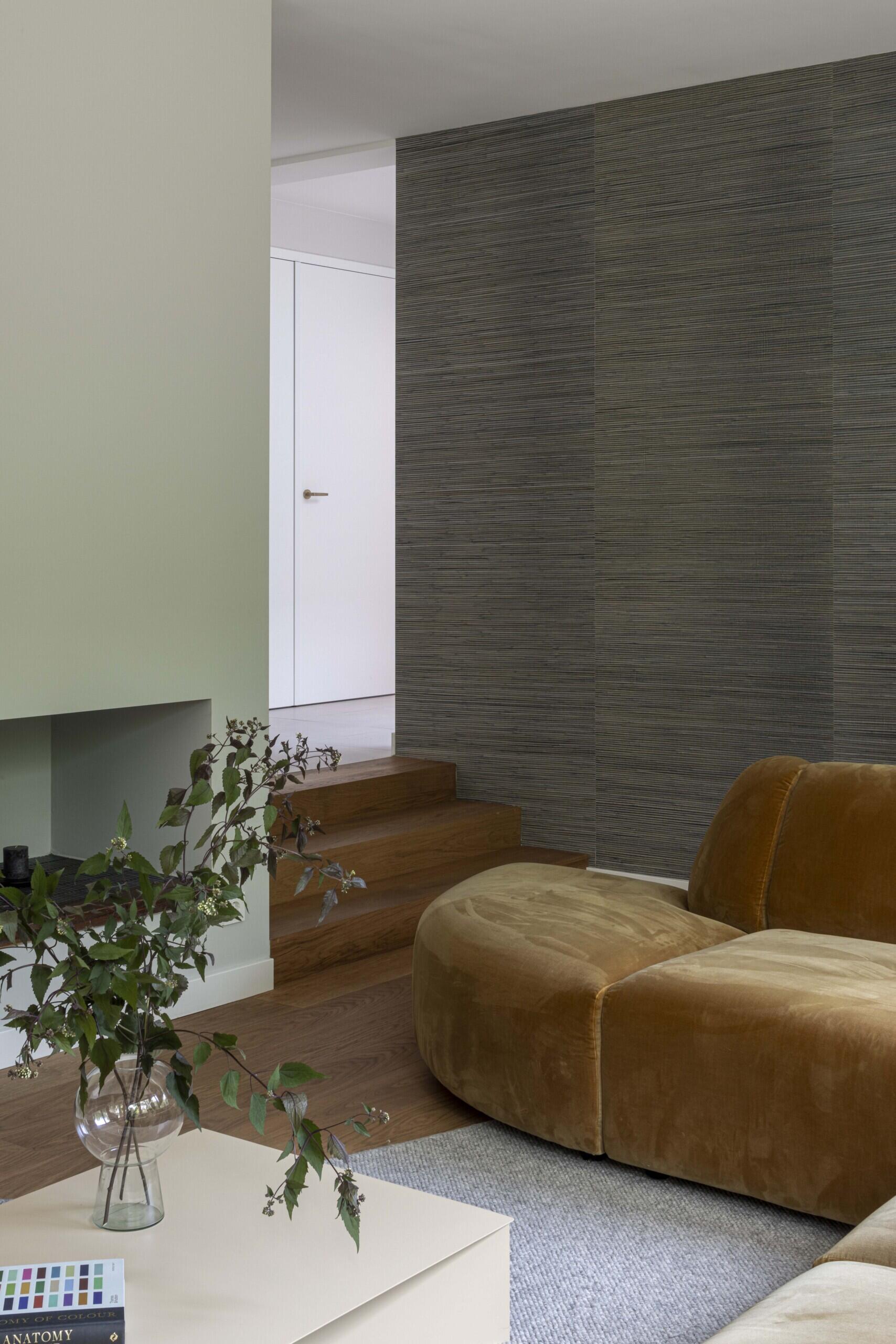 This living room blends comfort and calm with a strong sense of natural style. A large caramel-colored velvet sofa wraps around the space, offering a relaxed and sculptural focal point that invites conversation or quiet retreat.  The design favors earthy textures and muted tones, with warm wood flooring and a subtle gray wall finish that adds depth without distraction. A soft, curved rug defines the seating area, while a minimalist pale coffee table keeps the look light and open. Lush indoor plants echo the greenery visible through the full-height glass doors, creating a visual link to the garden just outside.  The result is a living space that feels warm, grounded, and thoughtfully connected to its surroundings.