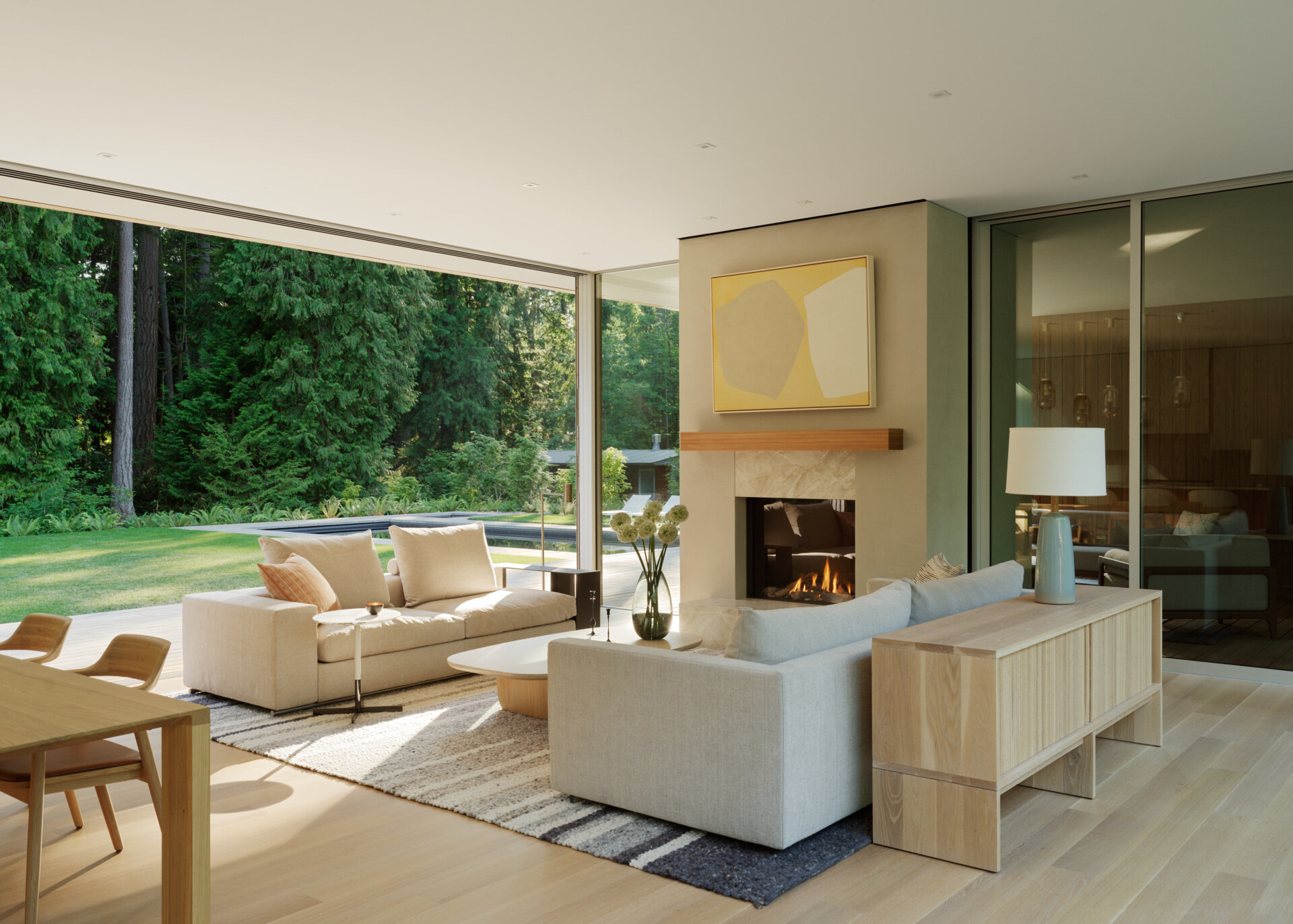 The living room is located in one of the long, linear wings and sits open to both the kitchen and dining areas. Generous glazing frames views of the surrounding trees, while high ceilings, a fireplace, and a restrained material palette give the space an easy, grounded elegance. Natural light is abundant, balanced across the day thanks to the wing’s narrow profile and window placement.