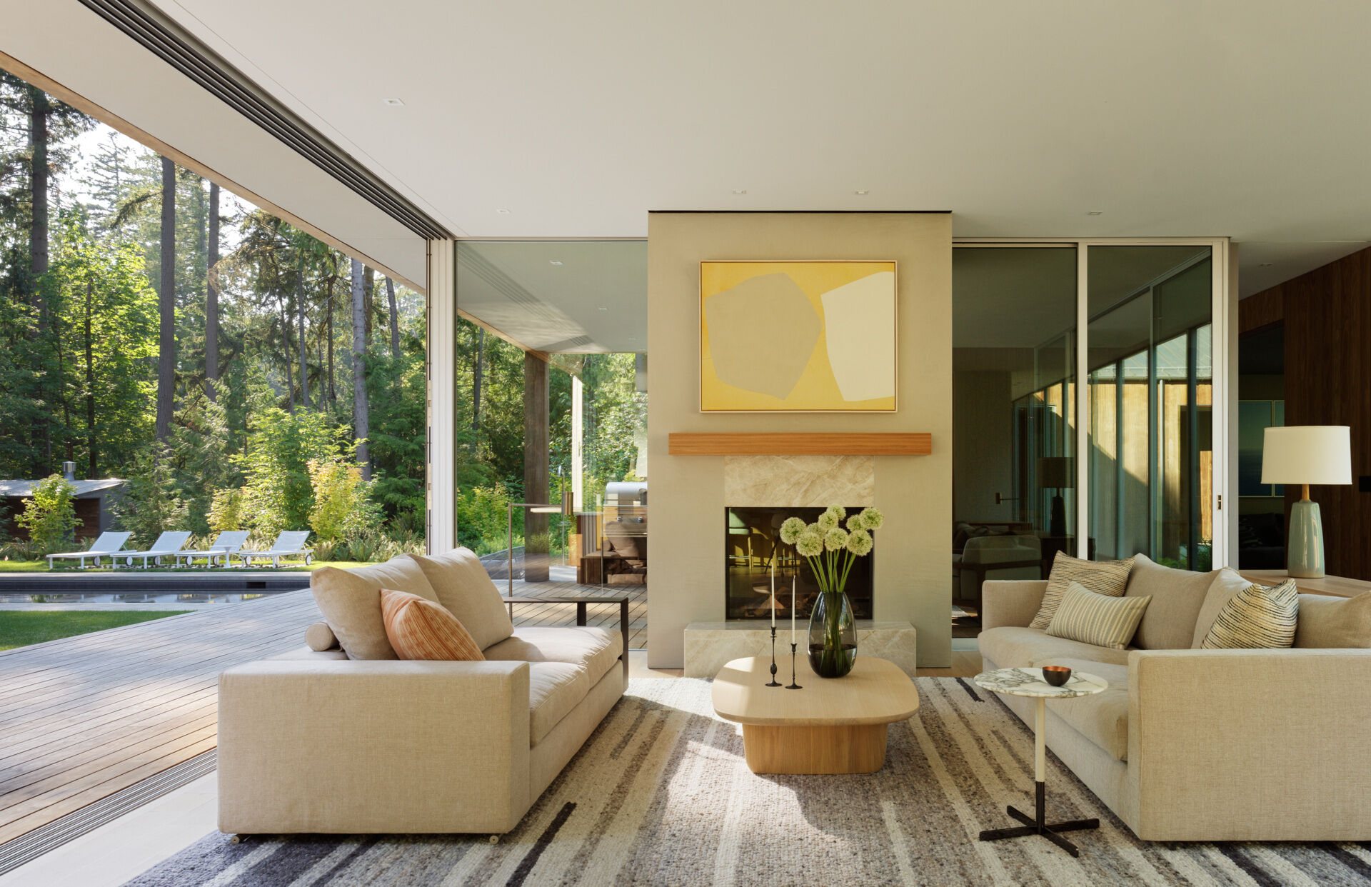 The living room is located in one of the long, linear wings and sits open to both the kitchen and dining areas. Generous glazing frames views of the surrounding trees, while high ceilings, a fireplace, and a restrained material palette give the space an easy, grounded elegance. Natural light is abundant, balanced across the day thanks to the wing’s narrow profile and window placement.