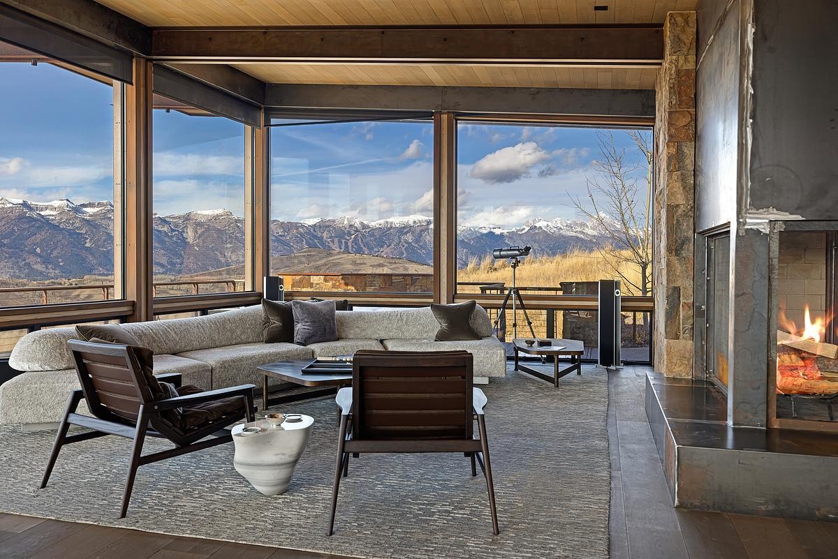 The exposed wood beams overhead lend a cozy sense of structure for the open plan interior, while soft textures in the rug and sectional invite you to sink in and stay awhile. Earth-toned pillows and sleek, sculptural chairs add just the right touch of contrast, while the fireplace, wrapped in stone and steel, creates a look that’s both grounded and modern.