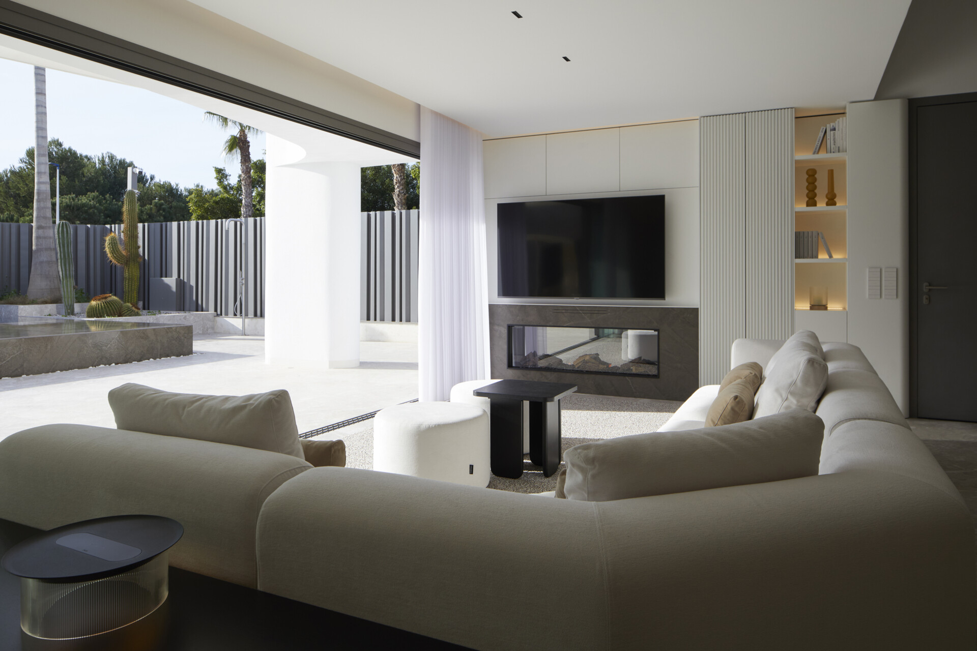 The living room opens wide to the outdoors, blurring the line between inside comfort and sunlit patio life. A soothing material palette of taupe porcelain, layered whites, and soft black and ochre touches sets the tone, while a sweeping sectional in a soft, sandy fabric echoes the curves of the exterior architecture. 

The fireplace, framed in a charcoal stone surround, anchors the room with understated elegance beneath the wall-mounted TV. Built-in shelves with warm backlighting display books and sculptural pieces, adding just the right touch of personality without feeling cluttered.
