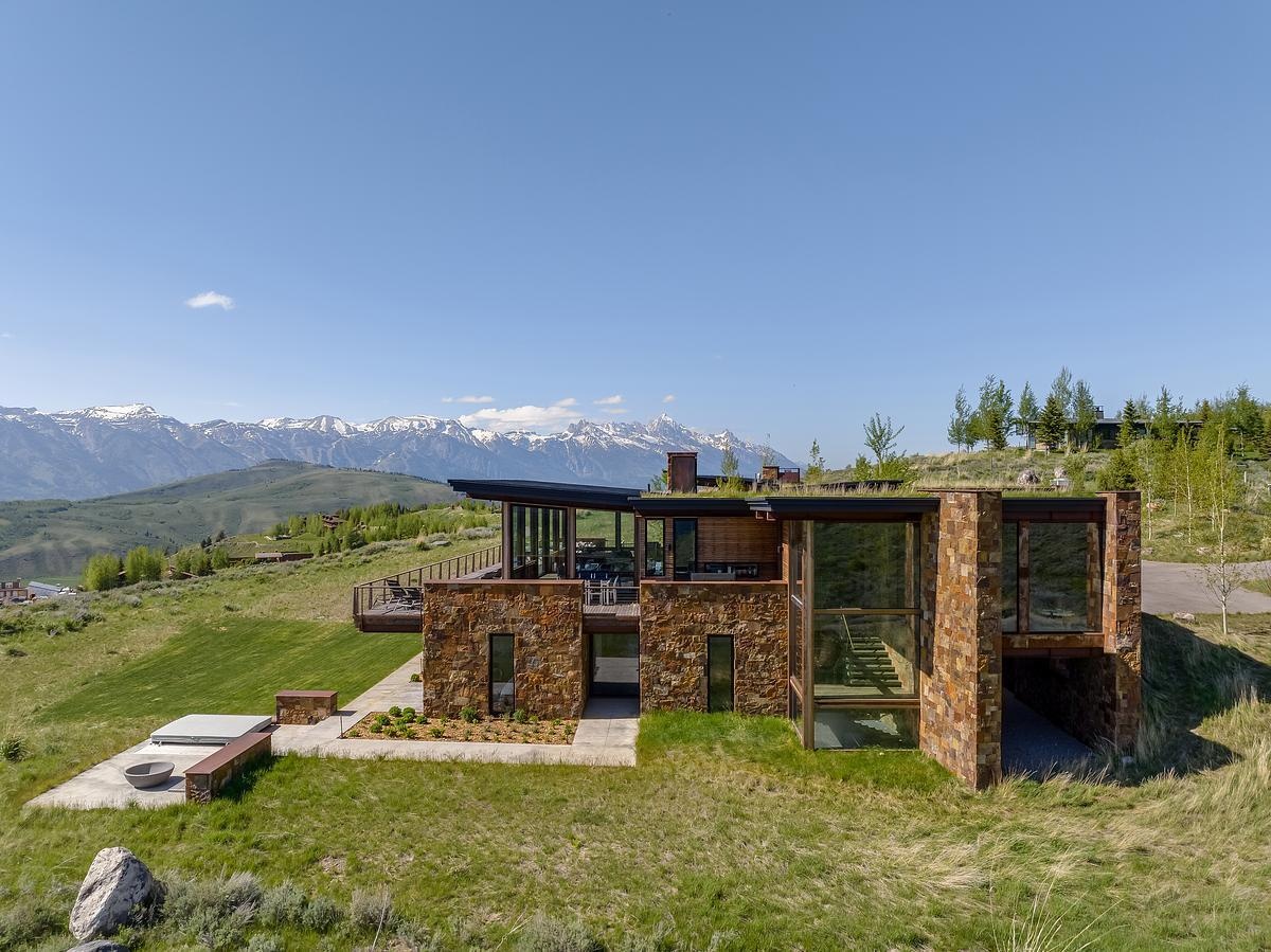 Nestled into the hillside like it naturally belongs there, this home embraces its mountain surroundings with a rugged elegance. The living roof, blanketed in grass, blends almost seamlessly into the wild landscape, while stacked stone walls ground the structure with earthy warmth. Generous windows invite the panoramic views inside, offering a daily dose of the snow-capped peaks beyond. It's the kind of place that feels both tucked away and wide open, perfect for quiet mornings or evenings that stretch out under the stars.
