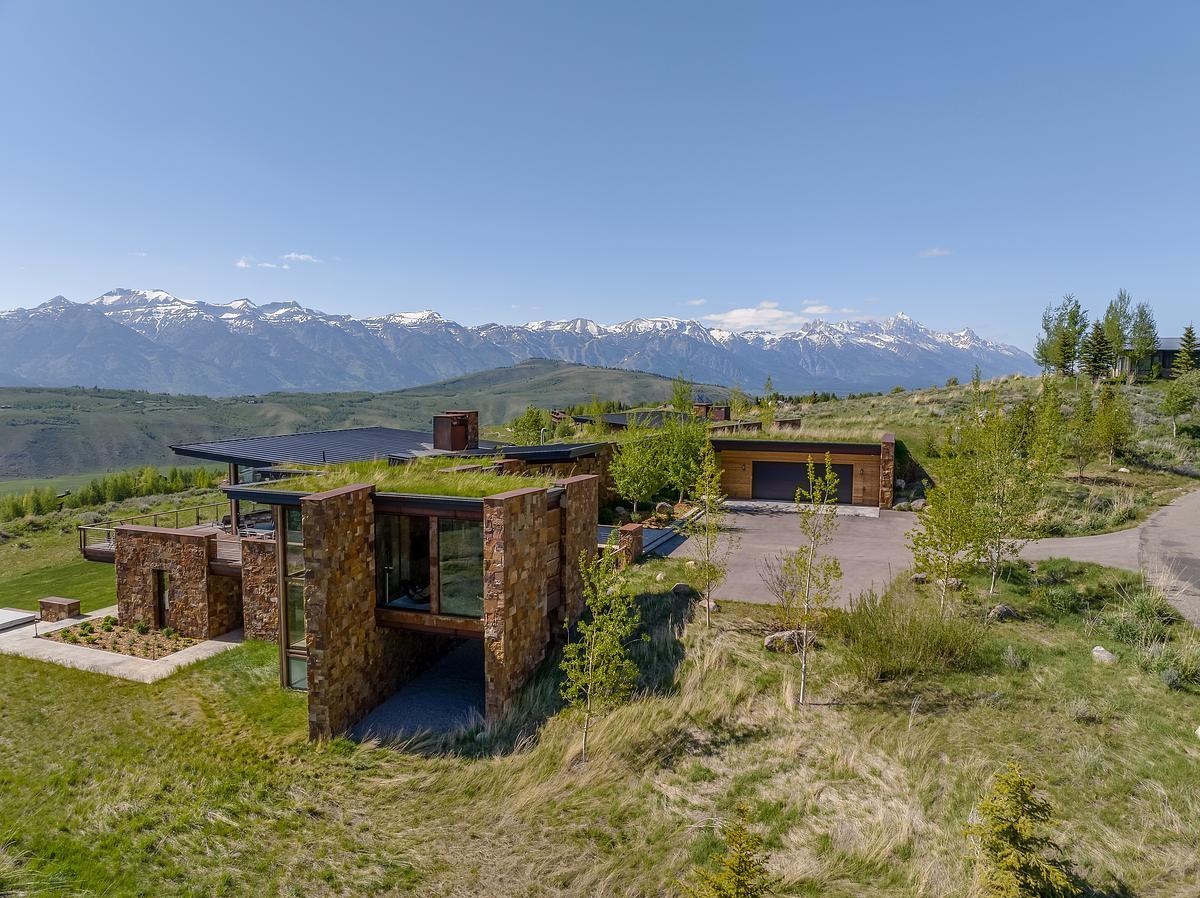 Nestled into the hillside like it naturally belongs there, this home embraces its mountain surroundings with a rugged elegance. The living roof, blanketed in grass, blends almost seamlessly into the wild landscape, while stacked stone walls ground the structure with earthy warmth. Generous windows invite the panoramic views inside, offering a daily dose of the snow-capped peaks beyond. It's the kind of place that feels both tucked away and wide open, perfect for quiet mornings or evenings that stretch out under the stars.