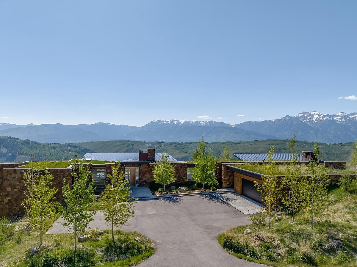 Nestled into the hillside like it naturally belongs there, this home embraces its mountain surroundings with a rugged elegance. The living roof, blanketed in grass, blends almost seamlessly into the wild landscape, while stacked stone walls ground the structure with earthy warmth. Generous windows invite the panoramic views inside, offering a daily dose of the snow-capped peaks beyond. It's the kind of place that feels both tucked away and wide open, perfect for quiet mornings or evenings that stretch out under the stars.