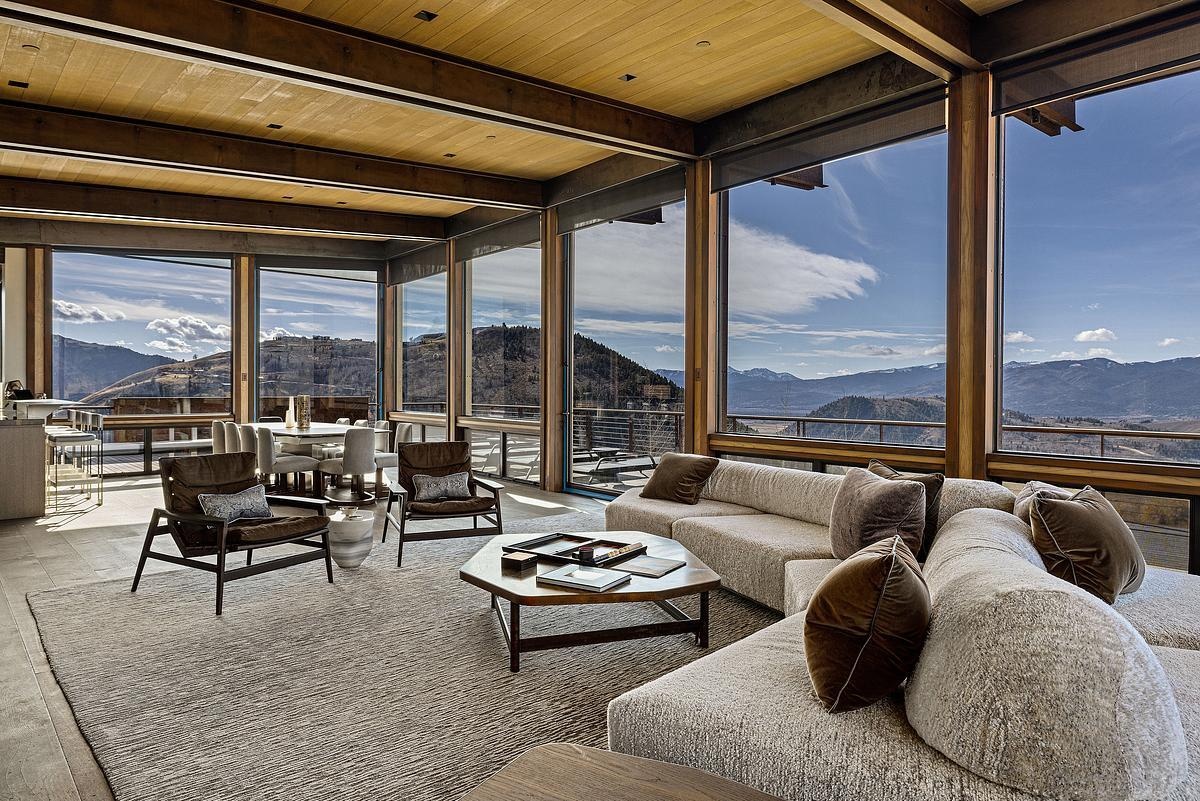 The exposed wood beams overhead lend a cozy sense of structure for the open plan interior, while soft textures in the rug and sectional invite you to sink in and stay awhile. Earth-toned pillows and sleek, sculptural chairs add just the right touch of contrast, while the fireplace, wrapped in stone and steel, creates a look that’s both grounded and modern.