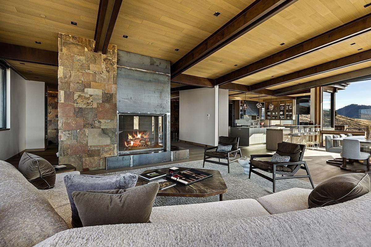 The exposed wood beams overhead lend a cozy sense of structure for the open plan interior, while soft textures in the rug and sectional invite you to sink in and stay awhile. Earth-toned pillows and sleek, sculptural chairs add just the right touch of contrast, while the fireplace, wrapped in stone and steel, creates a look that’s both grounded and modern.
