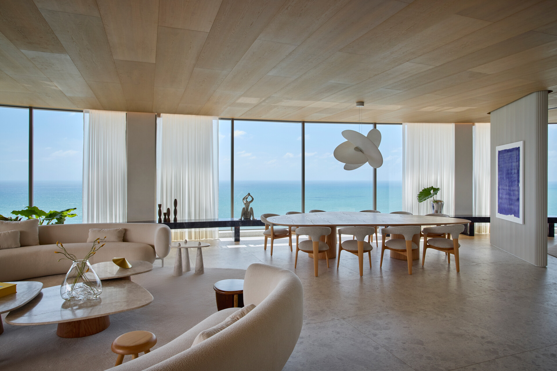 The dining area is part of an open-plan layout that includes the living room, kitchen, and barbecue area. Panels and natural stone bring warmth and texture, while soft, rounded furniture maintains a cohesive visual flow.