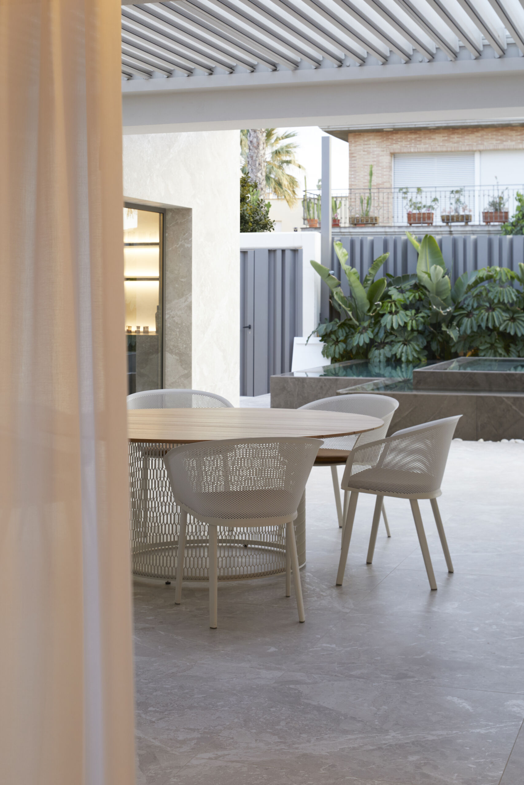 The covered outdoor dining space features circular wood table surrounded by chairs sits beneath a slatted pergola, offering filtered light and open-air comfort.