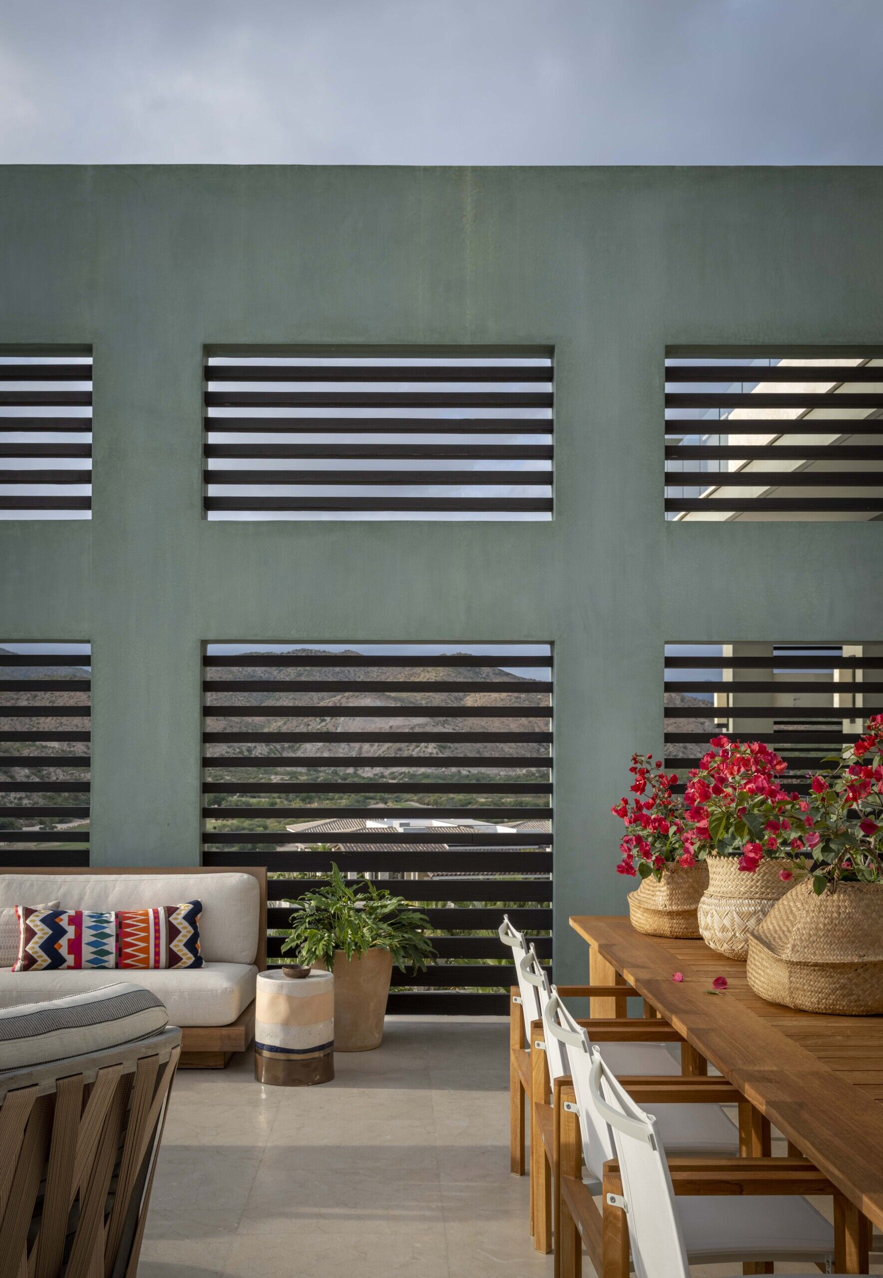 This outdoor space has a sage green wall with horizontal slats plays with light and shadow, letting in glimpses of the landscape beyond while still creating a cozy enclosure. A long wooden dining table anchors the space, topped with woven baskets overflowing with bright bougainvillea, their vivid pink petals offering a joyful burst of color. The white sling chairs keep the look fresh and breezy, perfect for lingering over a sunset meal.