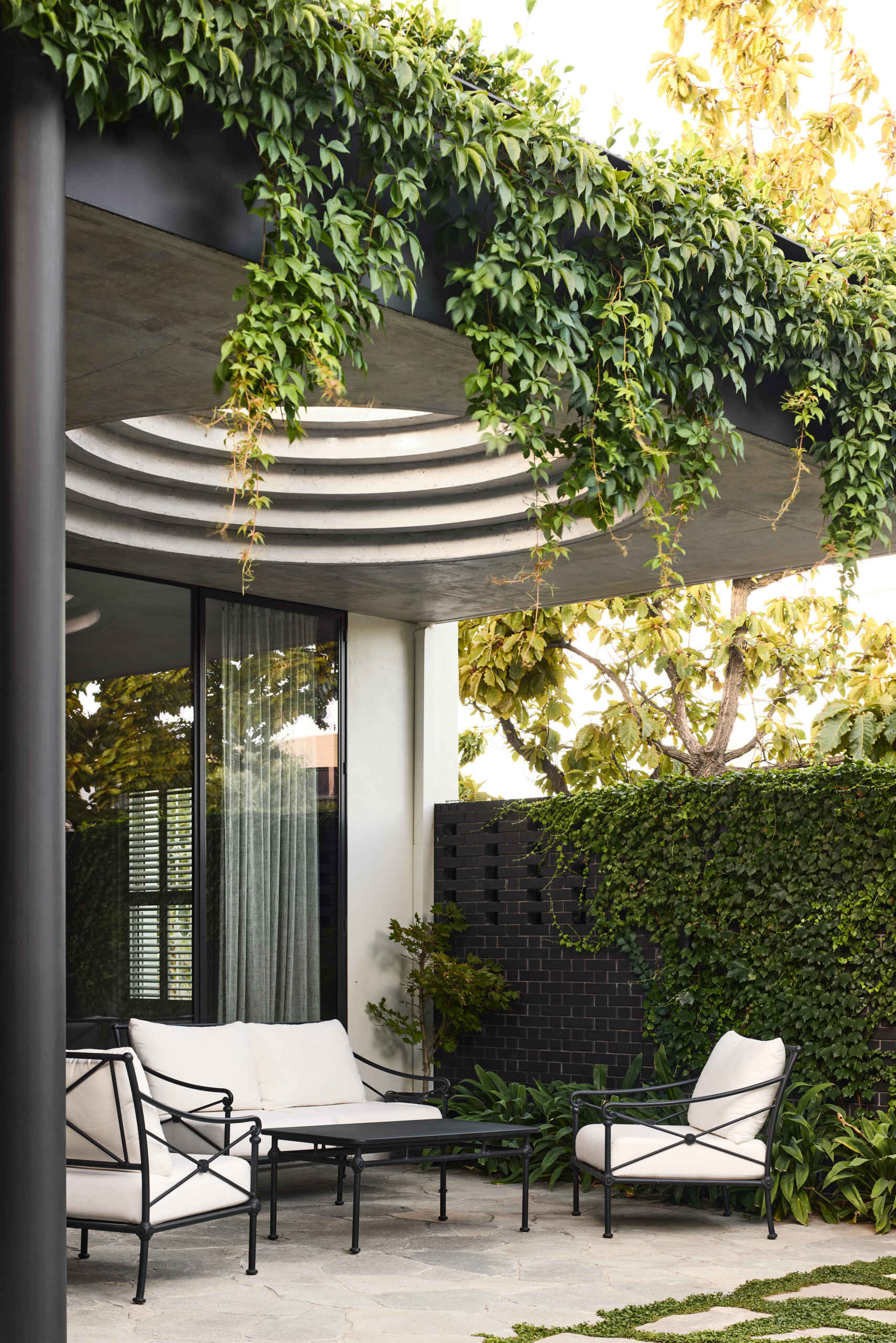 A defining feature of the rear of this remodeled home is a courtyard with a large circular cut-out overhead, a bold geometric move that invites sky views and dappled sunlight into the space below. Positioned above the outdoor dining area, the cut-out frames the moment, turning the patio into more than just a transitional space.