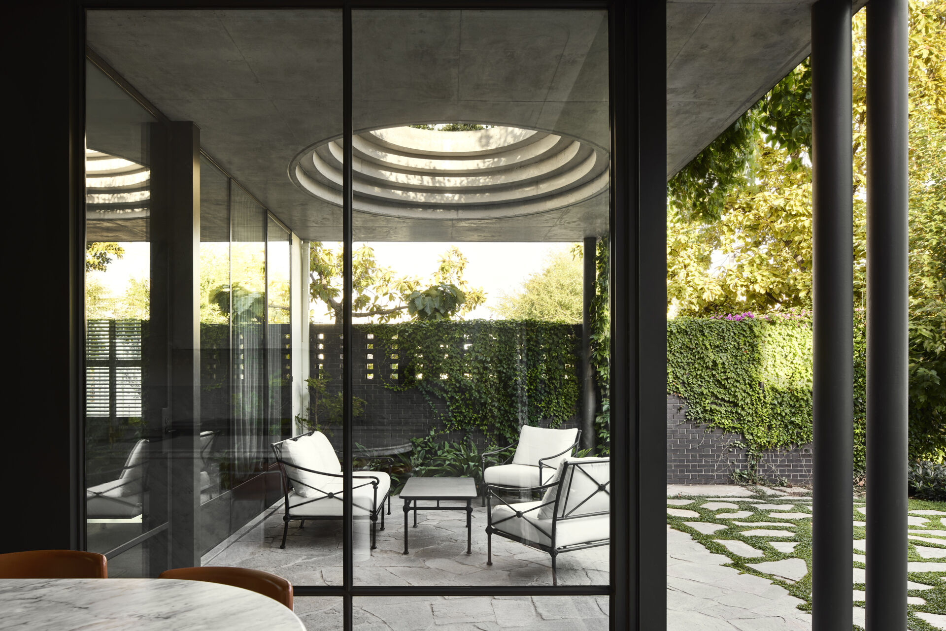 A defining feature of the rear of this remodeled home is a courtyard with a large circular cut-out overhead, a bold geometric move that invites sky views and dappled sunlight into the space below. Positioned above the outdoor dining area, the cut-out frames the moment, turning the patio into more than just a transitional space.