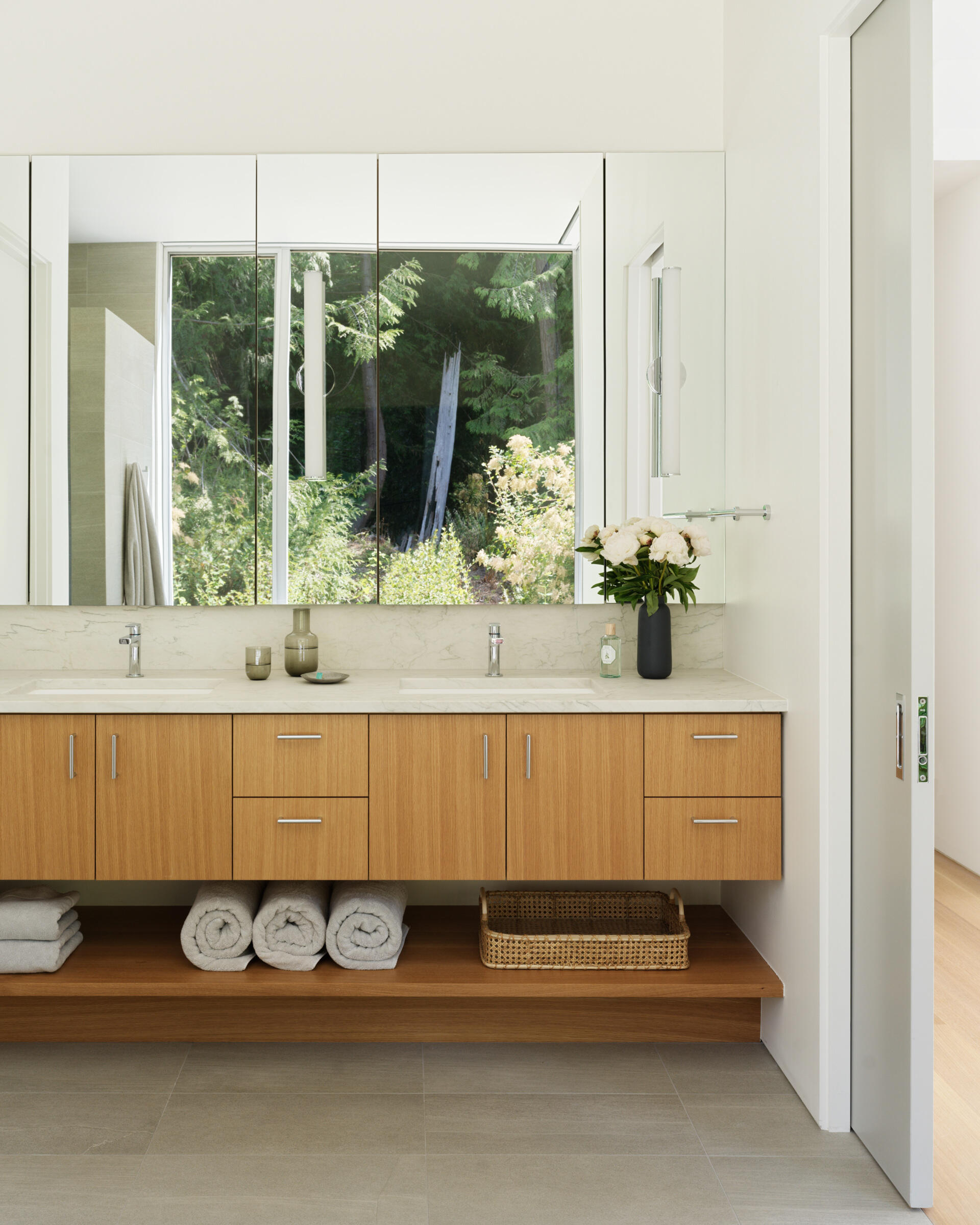 In the primary bath, impervious tile meets natural light in a calm, refined composition. A custom wood vanity brings warmth and texture to the room, with a streamlined design that includes an open shelf below for neatly stowed towels or baskets. Above, a large mirror reflects the light, expanding the sense of openness. 