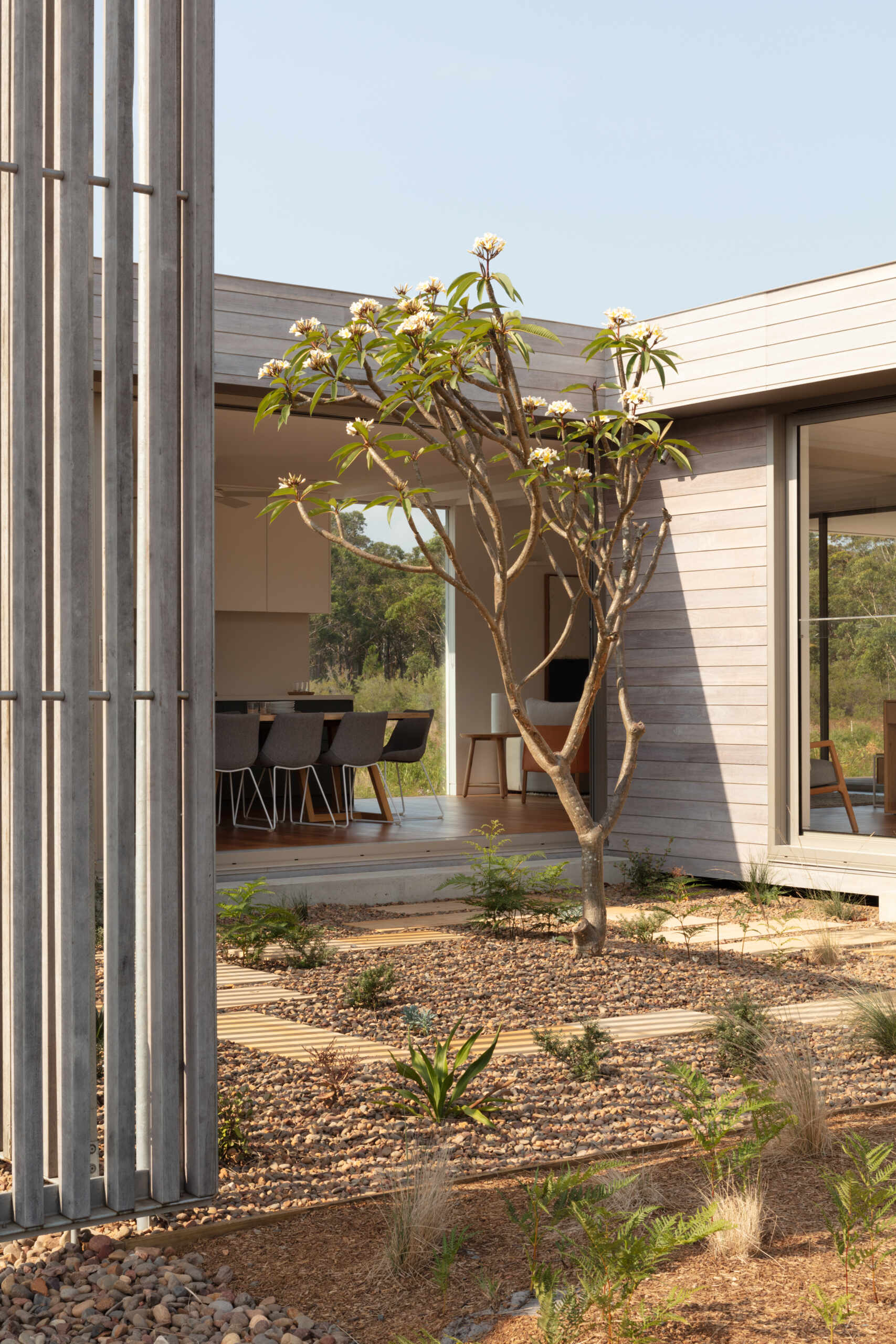 Pale wood slat fencing wraps the courtyard in a soft rhythm of vertical lines, offering privacy without blocking the light. A single tree reaches upward with graceful, sculptural limbs and fresh blossoms, adding just the right touch of life and color against the silvery backdrop. Below, a grid of sandstone pavers weaves through beds of pebbles and hardy greenery, creating a peaceful, low-maintenance oasis.