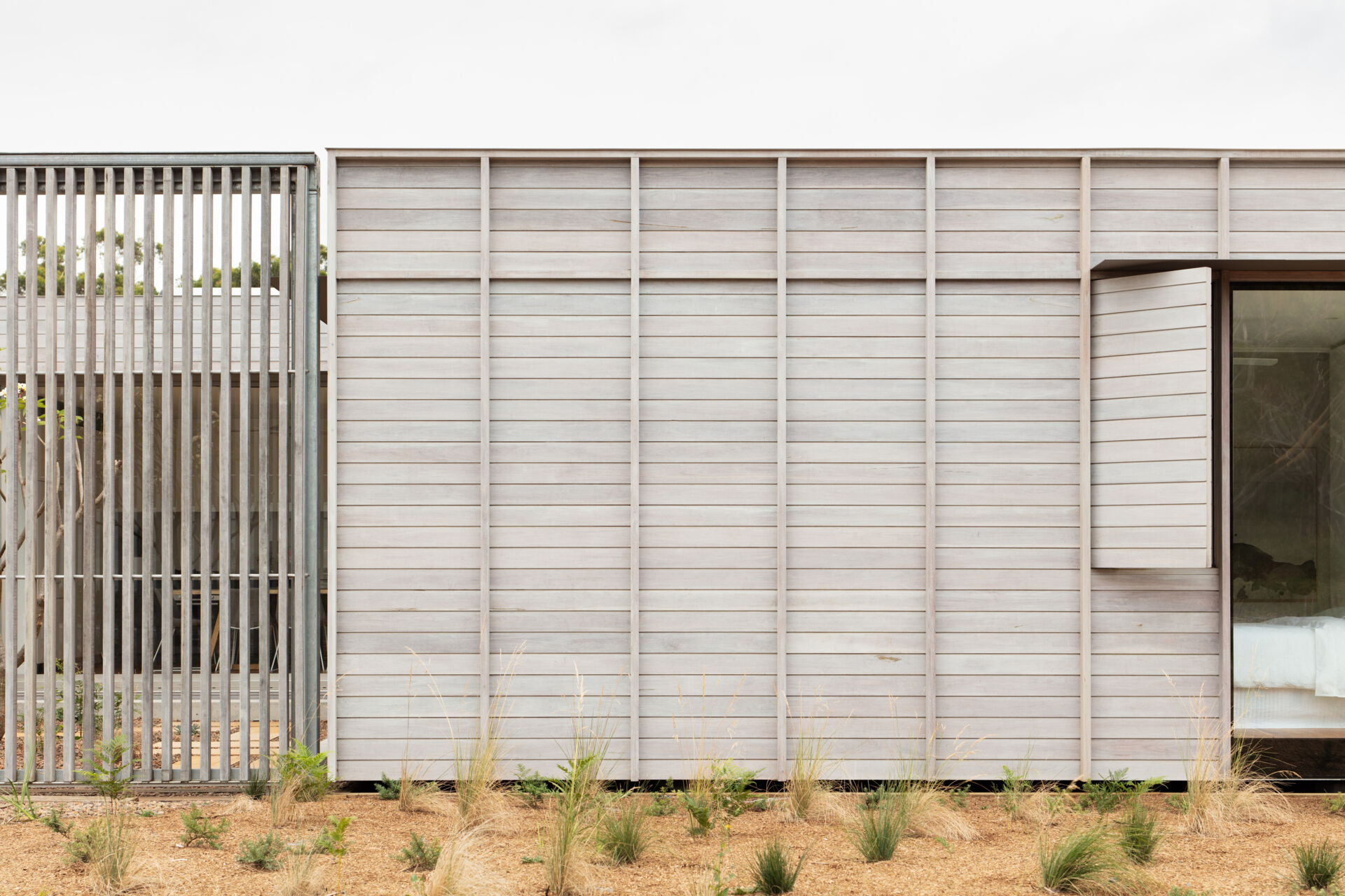 The pale timber cladding, with its subtle grain and muted tone, gives the facade a weathered elegance that blends beautifully into the sandy landscape. A clever shutter that opens like a page, reveals a narrow window beside a larger pane of glass.