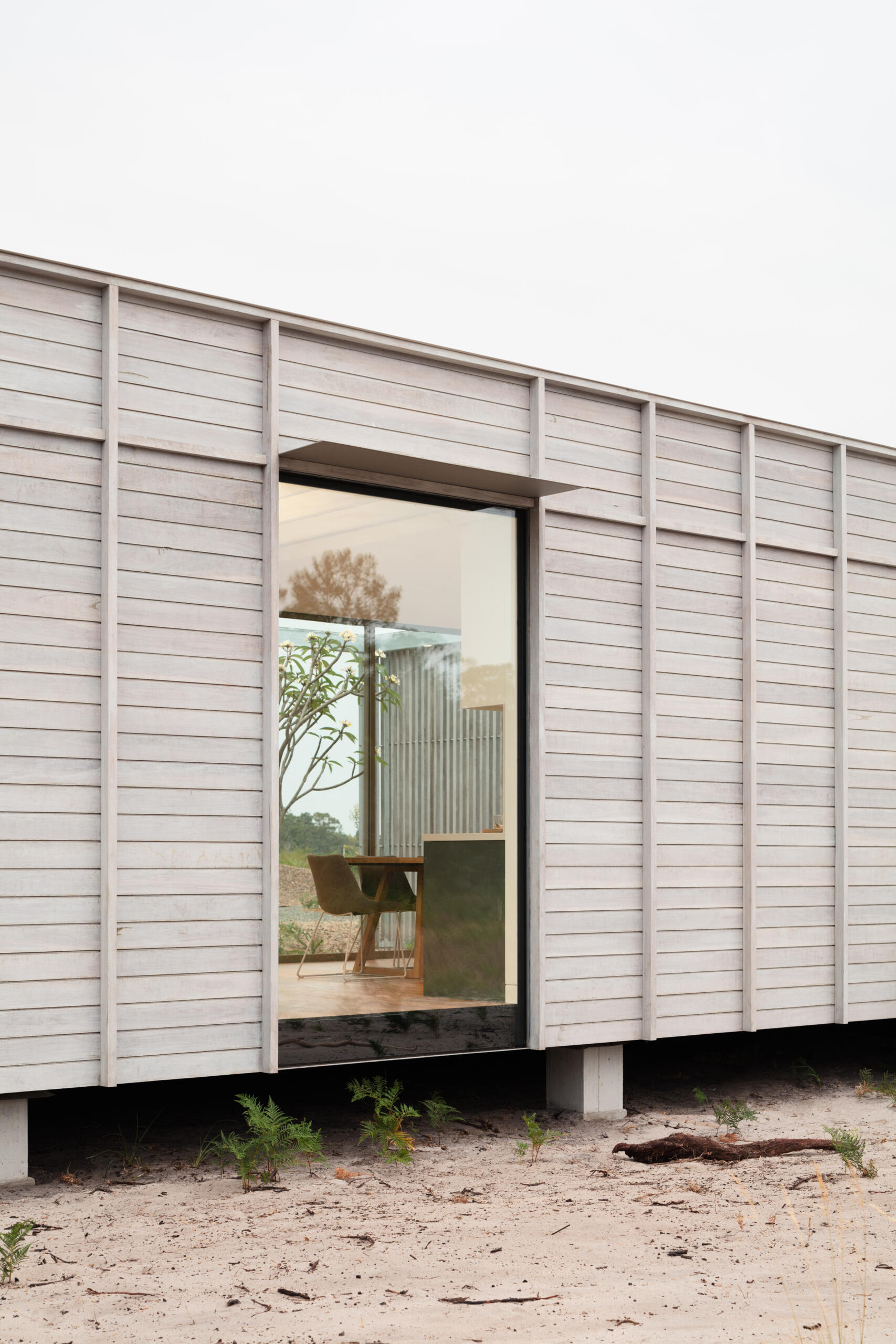 A large picture window punctuates the exterior, offering a view into the interior where a dining chair and table sit quietly, framed by glimpses of greenery beyond.