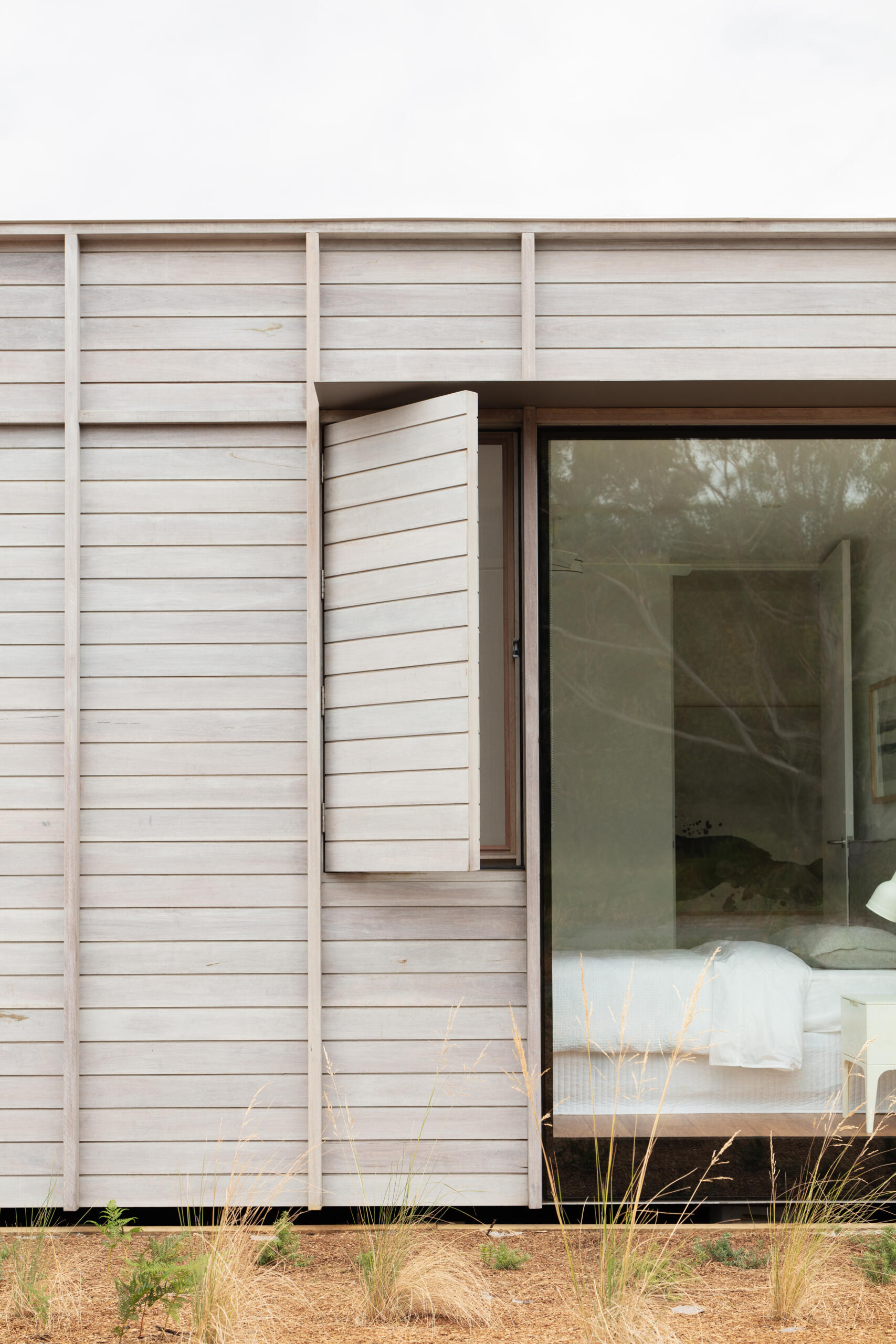 The pale timber cladding, with its subtle grain and muted tone, gives the facade a weathered elegance that blends beautifully into the sandy landscape. A clever shutter that opens like a page, reveals a narrow window beside a larger pane of glass.