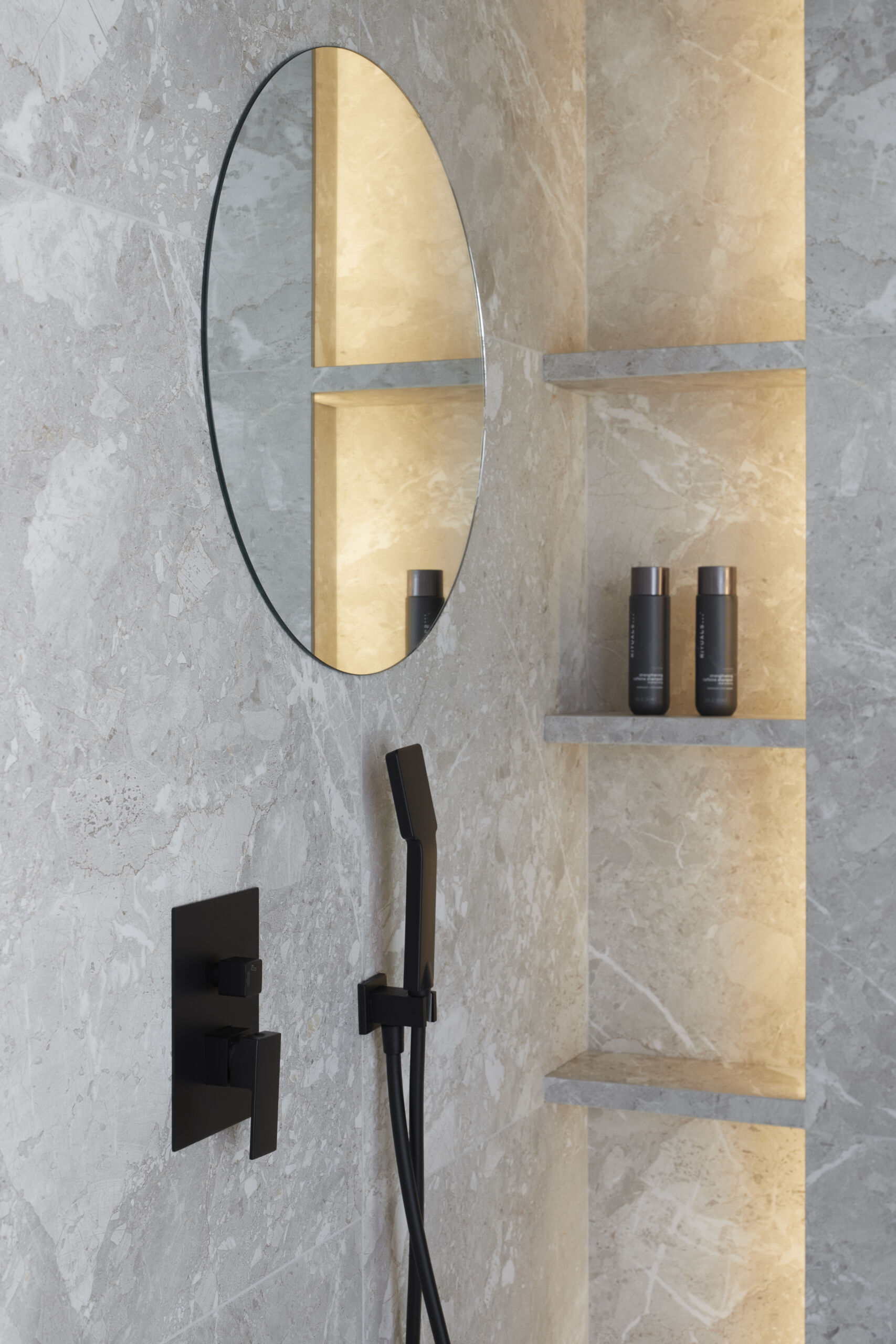 In the primary ensuite, a long stone vanity anchors this calming space, paired with softly glowing mirrors and matte black fixtures for contrast. Open shelves hold neatly folded towels, while a freestanding tub and black-lined recessed niche invite quiet, unhurried routines. Wrapped in soft gray stone, the shower pairs subtle texture with sharp black fixtures and a backlit niche. A frameless oval mirror adds a gentle curve, balancing the minimal palette with a quiet sense of care.