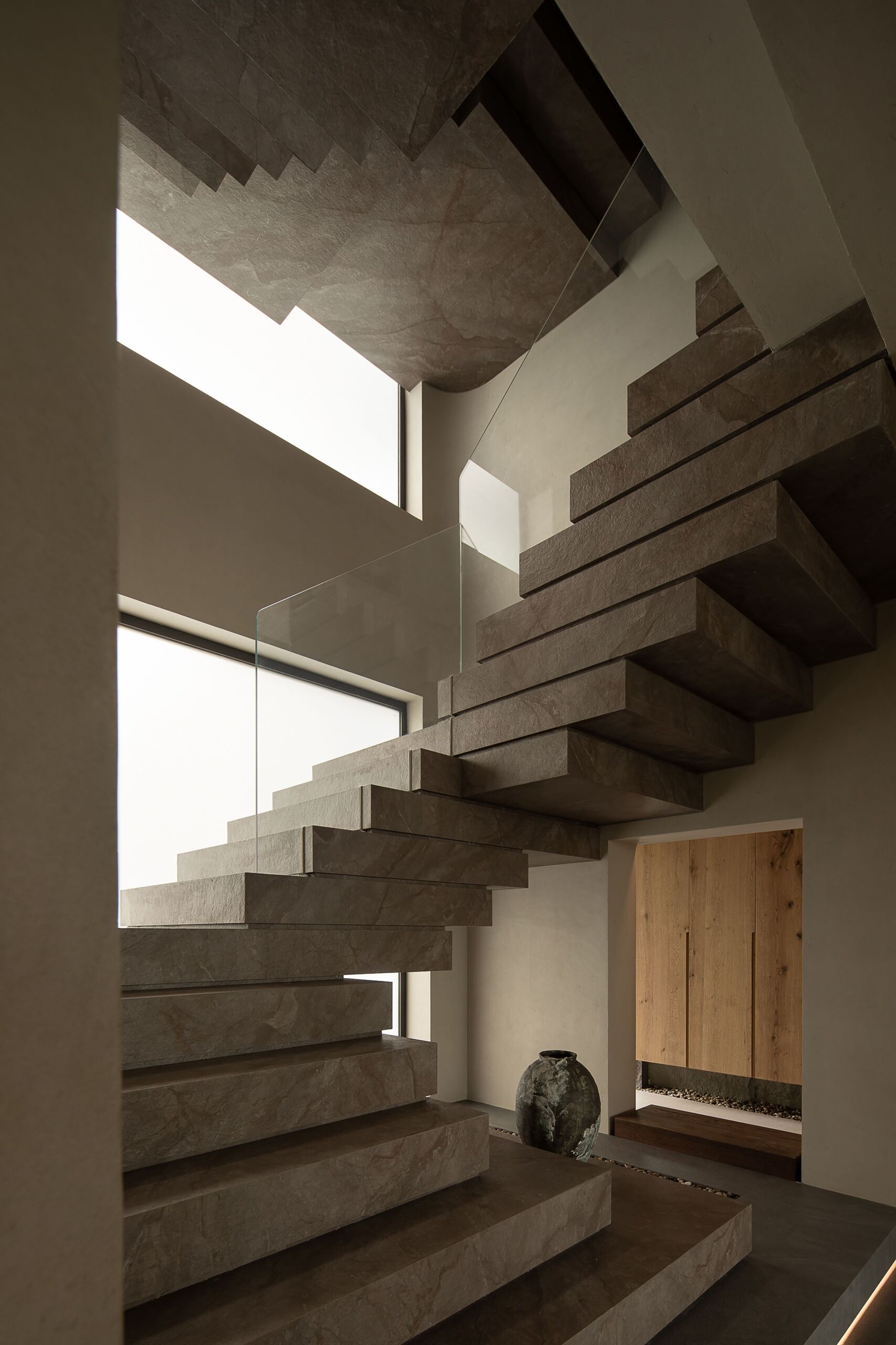 This sculptural staircase makes a bold architectural statement with its hefty stone treads and clean, floating form. Each step seems to hover in space, softened by the warm glow of daylight filtering through tall, frosted windows. A slim glass railing keeps the view uninterrupted, allowing the natural texture and subtle veining of the stone to shine.