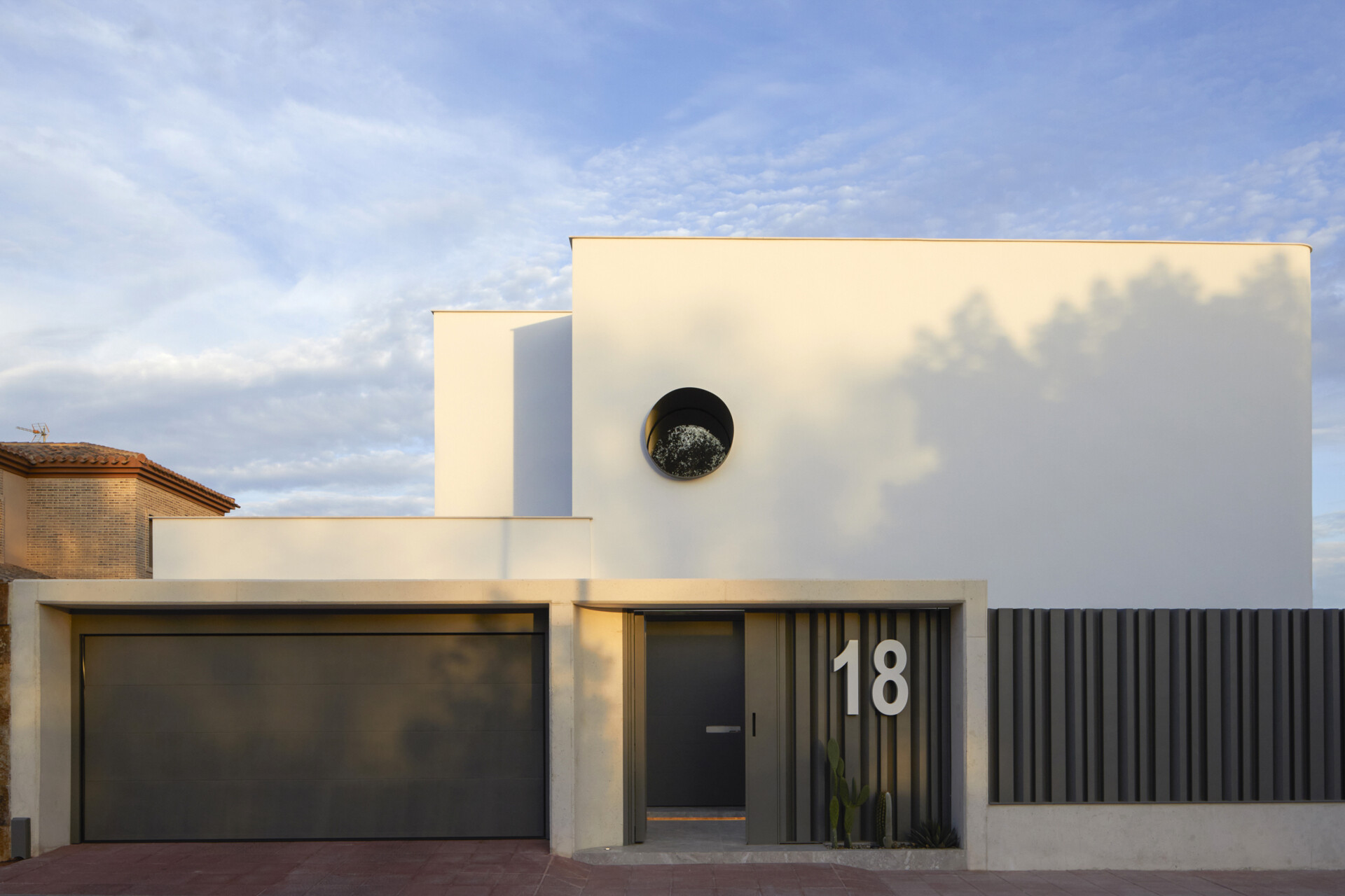 Bold House makes a strong first impression with its crisp white facade, softened by subtle curves and minimalist lines. A circular window punctuates the upper volume, offering a playful contrast to the otherwise rectilinear geometry. The street-facing entry is framed by textured vertical slats and oversized house numbers, setting the tone for the home’s bold yet balanced design language.