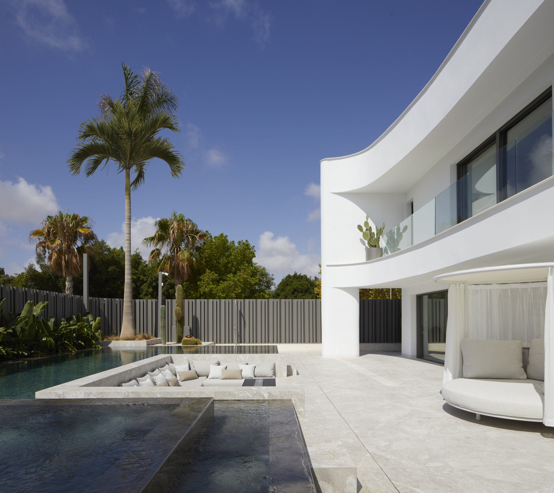 Recessed into the pool, the sunken lounge offers a sculpted retreat finished in pale stone. Deep built-in benches surround a sleek fire feature, while a plush arrangement of neutral-toned cushions adds comfort without disrupting the minimalist aesthetic.