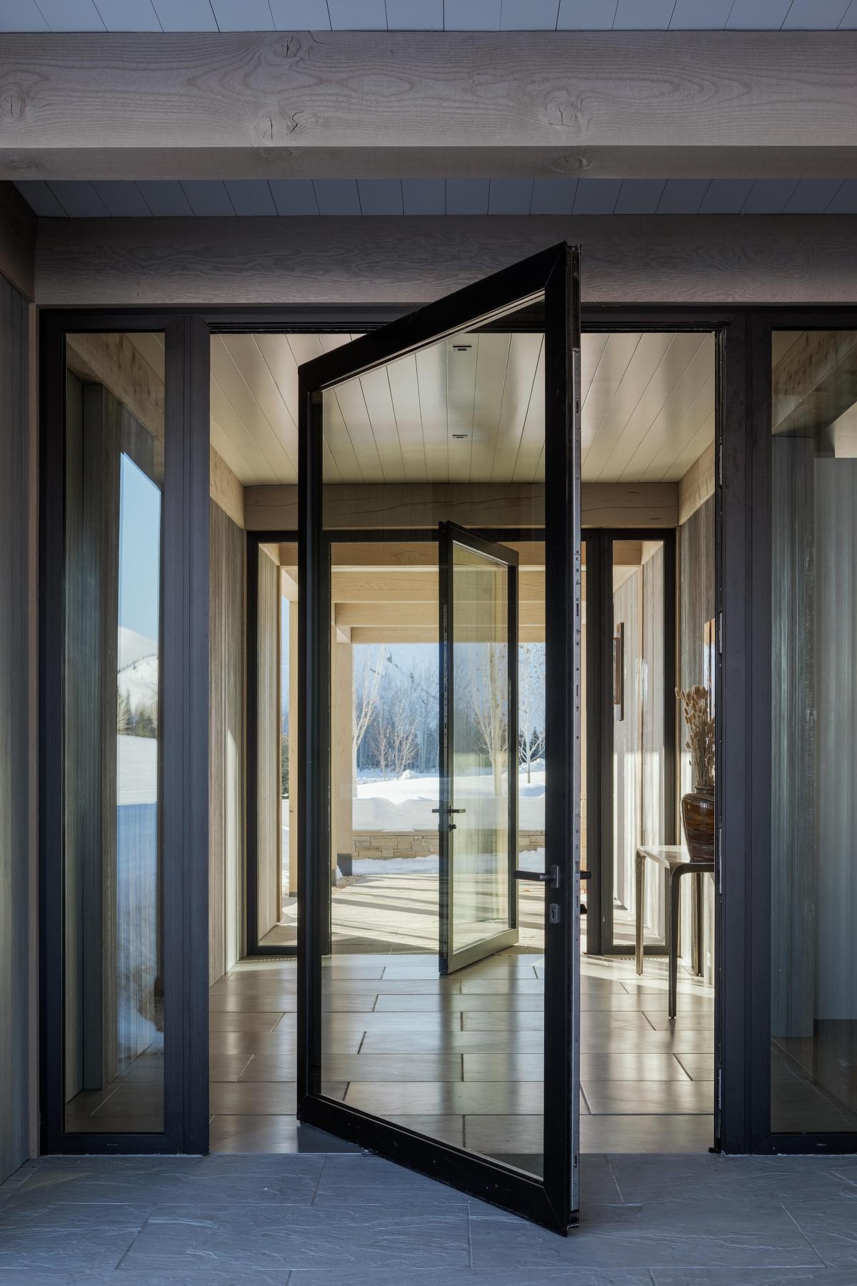 A breezeway entrance feels equal parts welcoming and intentional, with its light wood beams, soft stonework, pivoting glass doors, and clean-lined architecture drawing your eye straight through to the landscape beyond.
