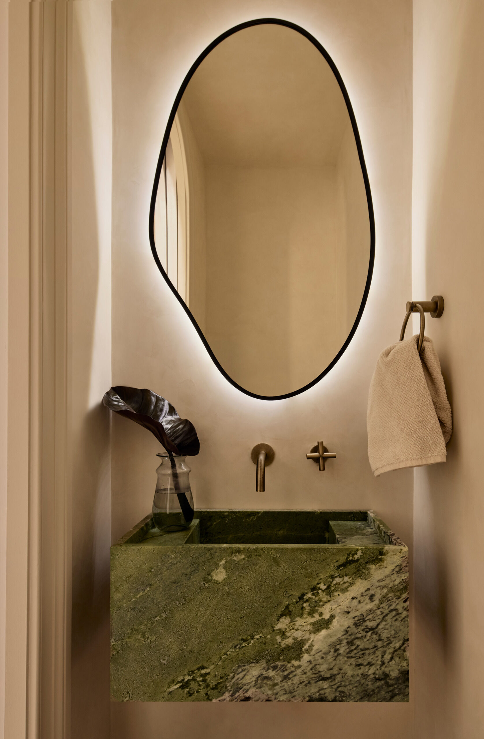 Compact yet luxurious, this powder room features creamy plaster walls and a sculptural Irish Connemara marble sink. A backlit asymmetrical mirror adds dimension and softness to the narrow space.