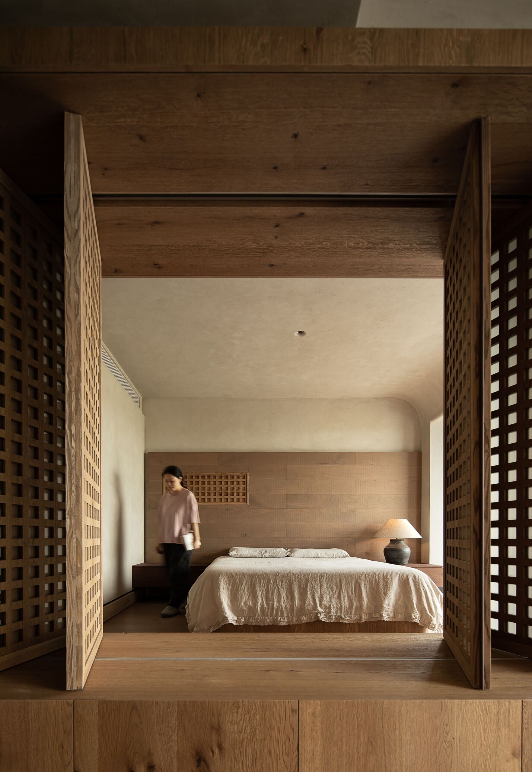 This primary bedroom feels like a peaceful retreat wrapped in wood and softness. Sliding lattice panels frame the space with a quiet rhythm, offering both privacy and texture. The bed sits low and grounded, dressed in a natural fabric cover that echoes the relaxed tone of the room. Warm light from a pottery-style lamp glows gently against the wood-paneled wall, while soft curves in the ceiling and corners create a cocoon-like atmosphere.
