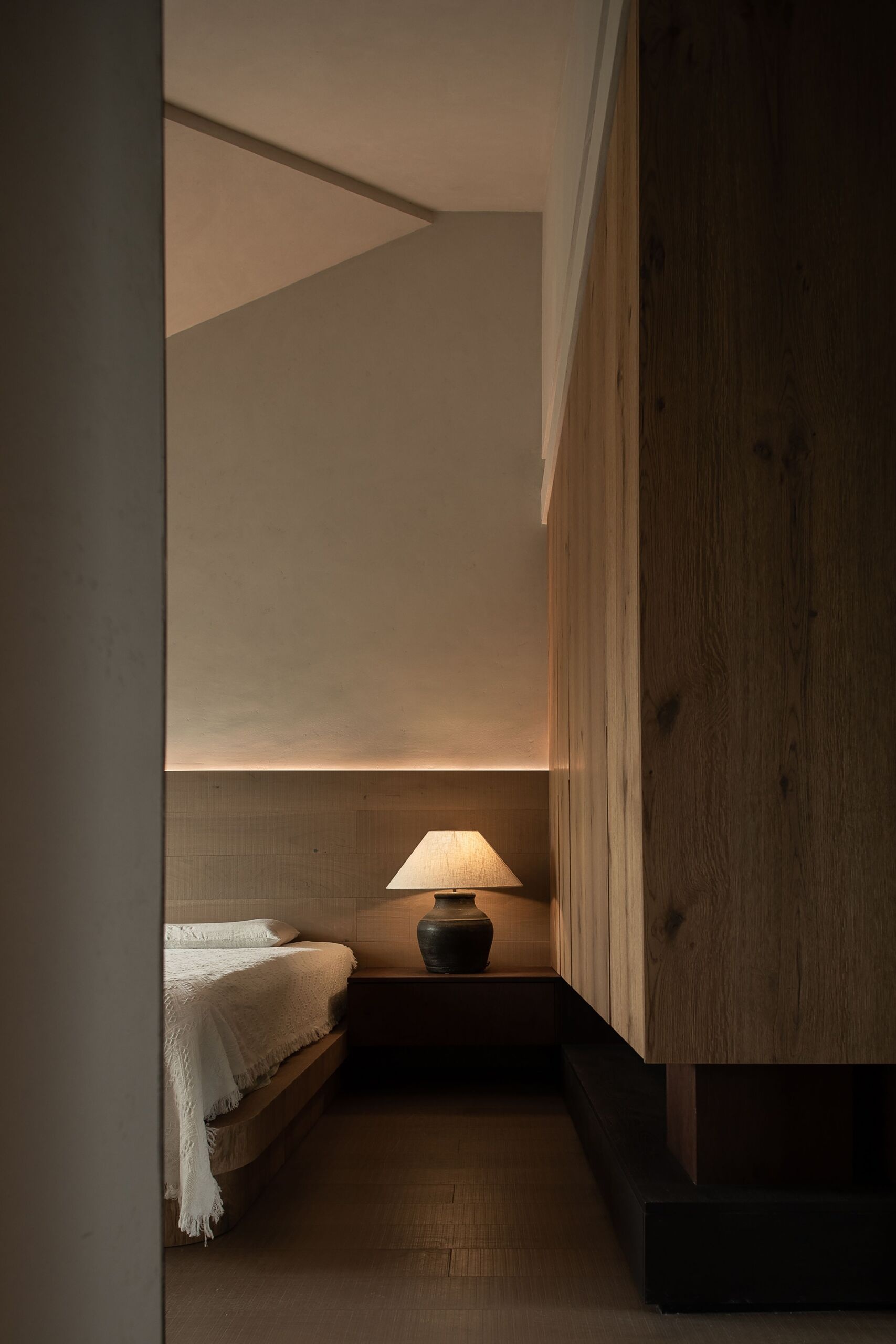 This primary bedroom feels like a peaceful retreat wrapped in wood and softness. Sliding lattice panels frame the space with a quiet rhythm, offering both privacy and texture. The bed sits low and grounded, dressed in a natural fabric cover that echoes the relaxed tone of the room. Warm light from a pottery-style lamp glows gently against the wood-paneled wall, while soft curves in the ceiling and corners create a cocoon-like atmosphere.