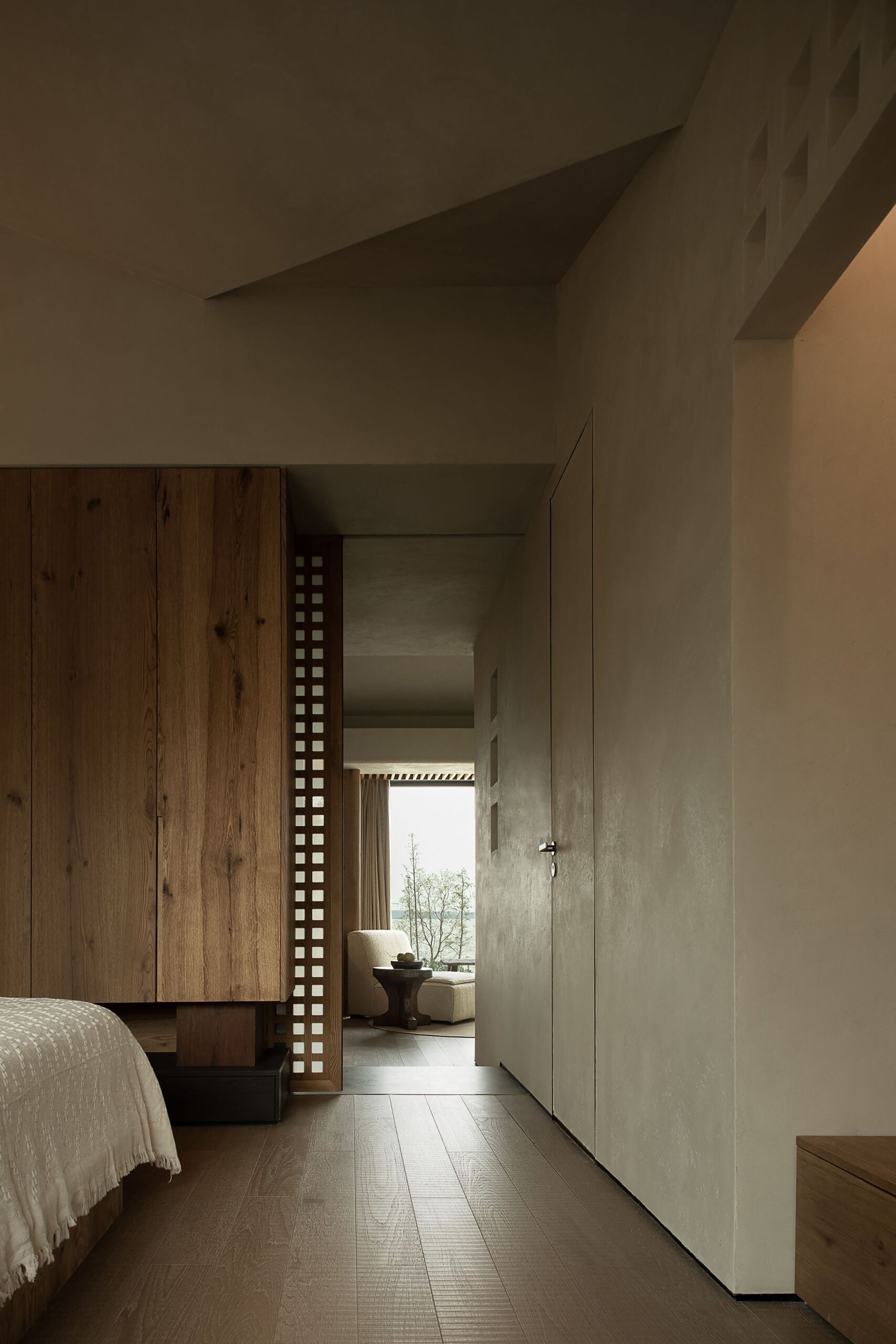 This primary bedroom feels like a peaceful retreat wrapped in wood and softness. Sliding lattice panels frame the space with a quiet rhythm, offering both privacy and texture. The bed sits low and grounded, dressed in a natural fabric cover that echoes the relaxed tone of the room. Warm light from a pottery-style lamp glows gently against the wood-paneled wall, while soft curves in the ceiling and corners create a cocoon-like atmosphere.