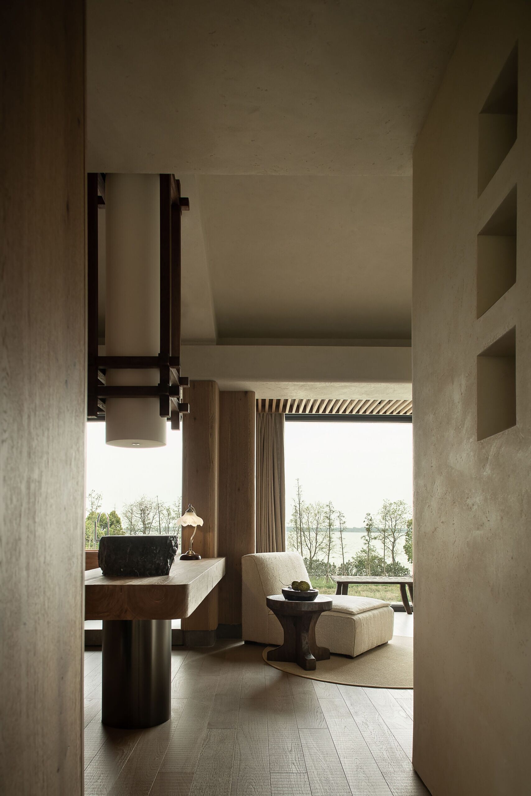This calming corner in the primary suite strikes a rare balance between work and rest. A sculpted wood desk stretches beneath a high ceiling, grounded by quiet tones and soft textures. Natural light pours in through floor-to-ceiling glass, opening the space to a tranquil garden view. Just beyond, a curved wood bathtub sits gently on a raised platform, inviting slow soaks after focused hours. With its layered materials, vintage-style lamp, and serene outlook, the room feels thoughtful, warm, and deeply connected to the outdoors.