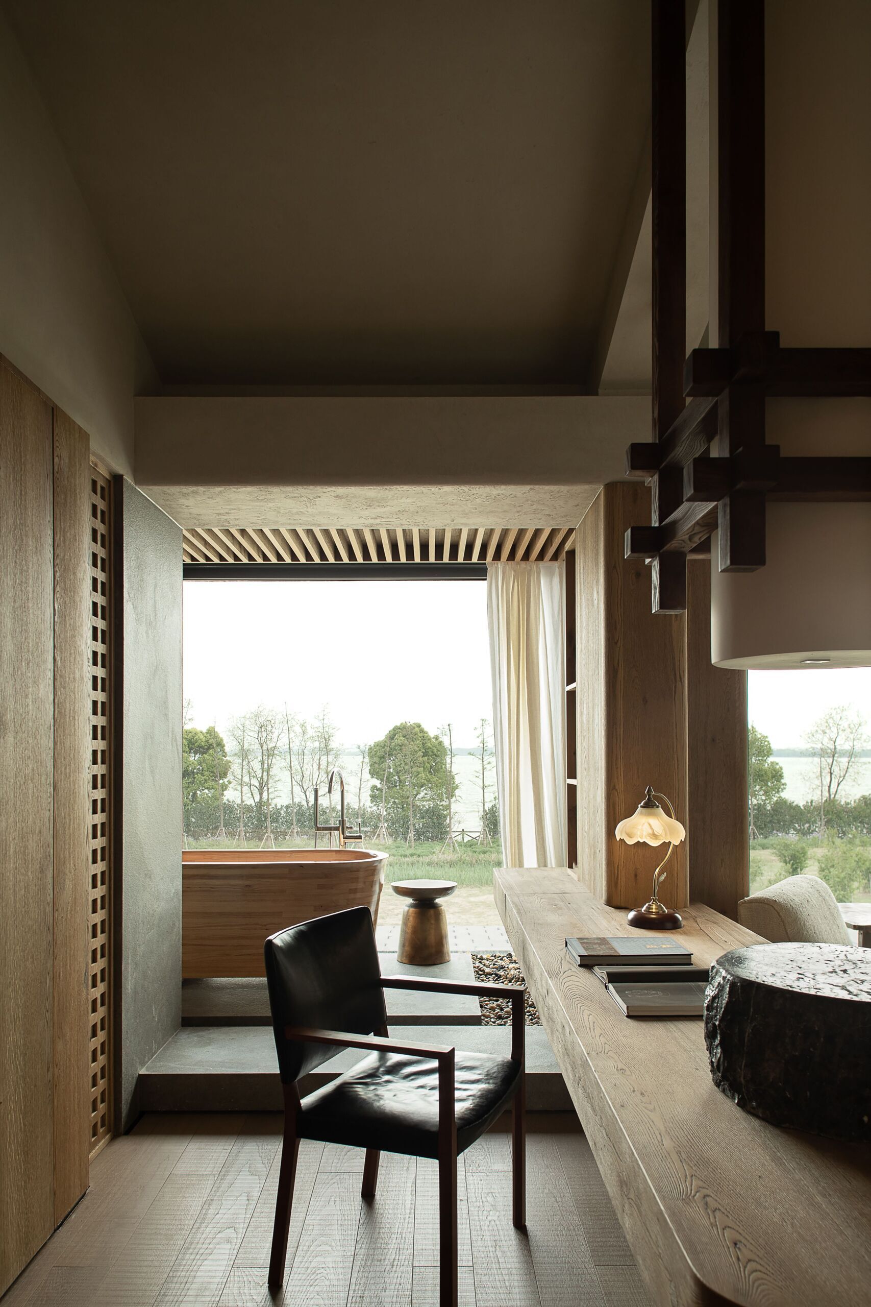 This calming corner in the primary suite strikes a rare balance between work and rest. A sculpted wood desk stretches beneath a high ceiling, grounded by quiet tones and soft textures. Natural light pours in through floor-to-ceiling glass, opening the space to a tranquil garden view. Just beyond, a curved wood bathtub sits gently on a raised platform, inviting slow soaks after focused hours. With its layered materials, vintage-style lamp, and serene outlook, the room feels thoughtful, warm, and deeply connected to the outdoors.
