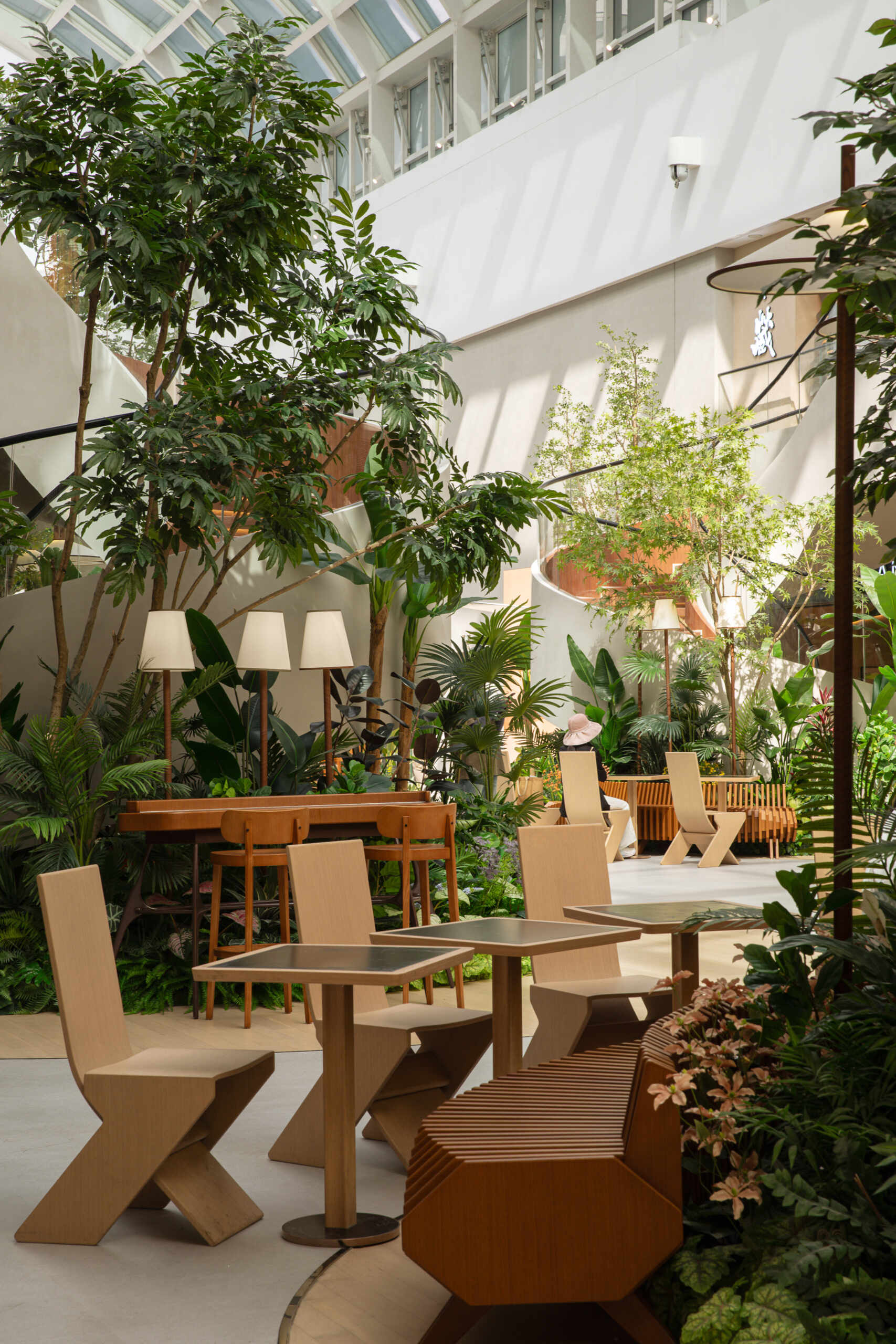 In this public plaza, long benches curve around clusters of greenery while sculptural chairs create relaxed, flexible seating. This dining space flows into a shared public garden, dissolving the line between restaurant and rest stop.