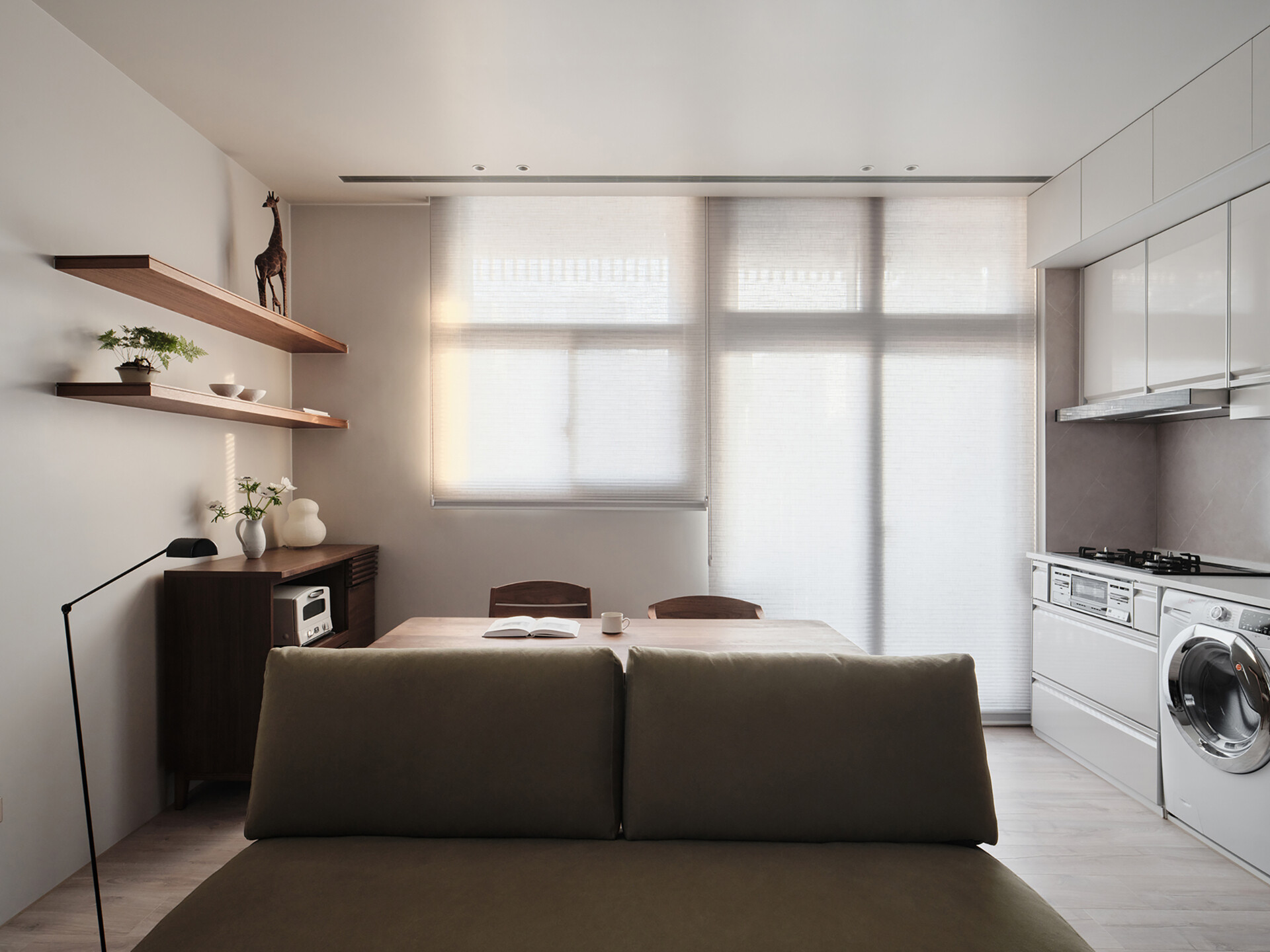 Soft morning light filters through sheer blinds, casting a gentle glow across this cozy, multi-functional space. The olive-toned sofa faces a tidy kitchen and dining area, where warm wood tones and clean white cabinetry balance comfort with modern efficiency. Floating shelves display just a few carefully chosen pieces, a bonsai, ceramics, and a charming giraffe, adding personality without clutter.