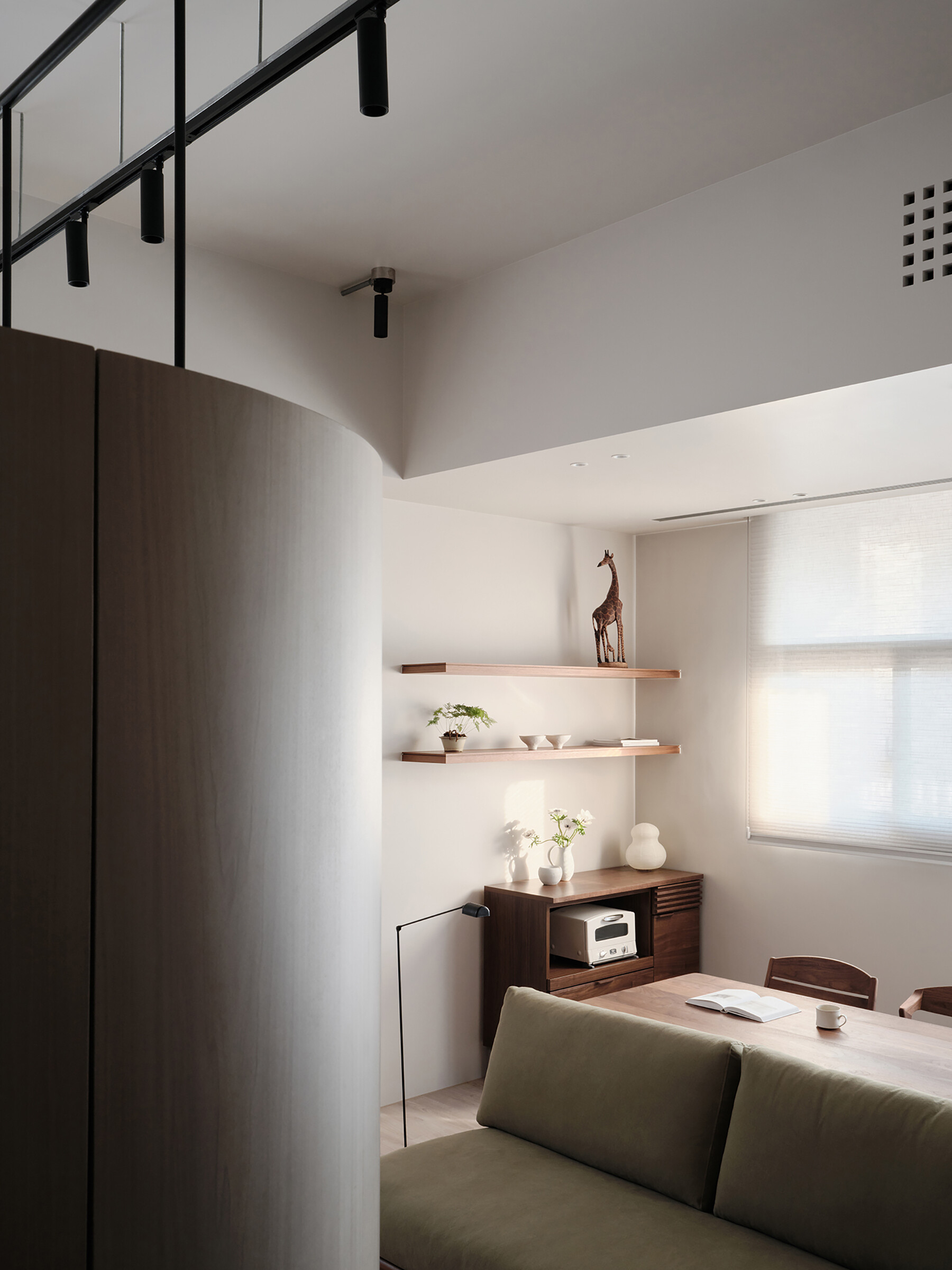 Soft morning light filters through sheer blinds, casting a gentle glow across this cozy, multi-functional space. The olive-toned sofa faces a tidy kitchen and dining area, where warm wood tones and clean white cabinetry balance comfort with modern efficiency. Floating shelves display just a few carefully chosen pieces, a bonsai, ceramics, and a charming giraffe, adding personality without clutter.
