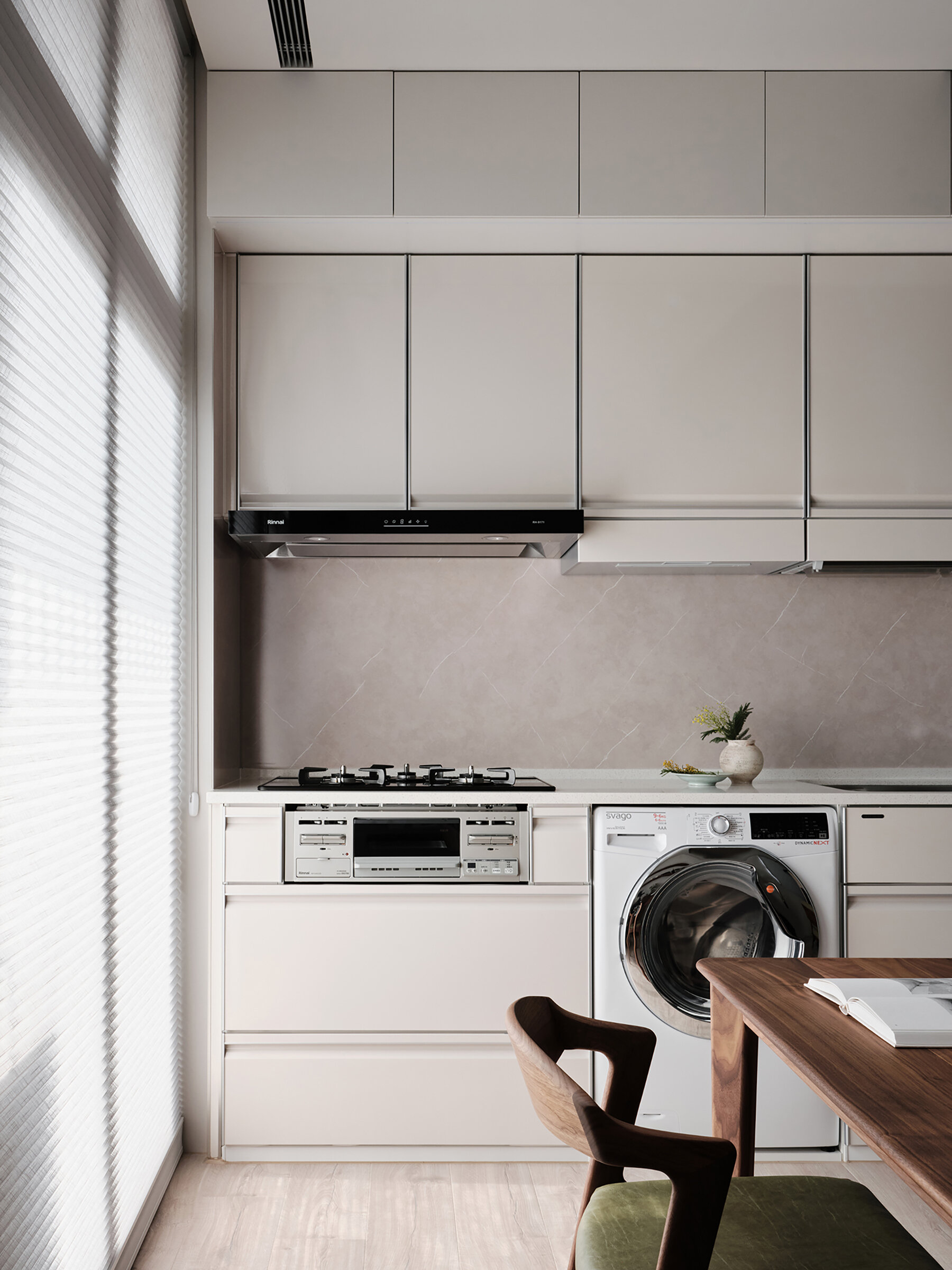 This compact kitchen feels effortlessly refined, with sleek cabinetry in a soft, warm beige that blends seamlessly into the walls. A subtle stone-look backsplash and pale wood flooring keep the palette light and harmonious. Built-in appliances, including a cooktop and washing machine, are tucked neatly into the lower cabinets, maximizing functionality without disrupting the flow.