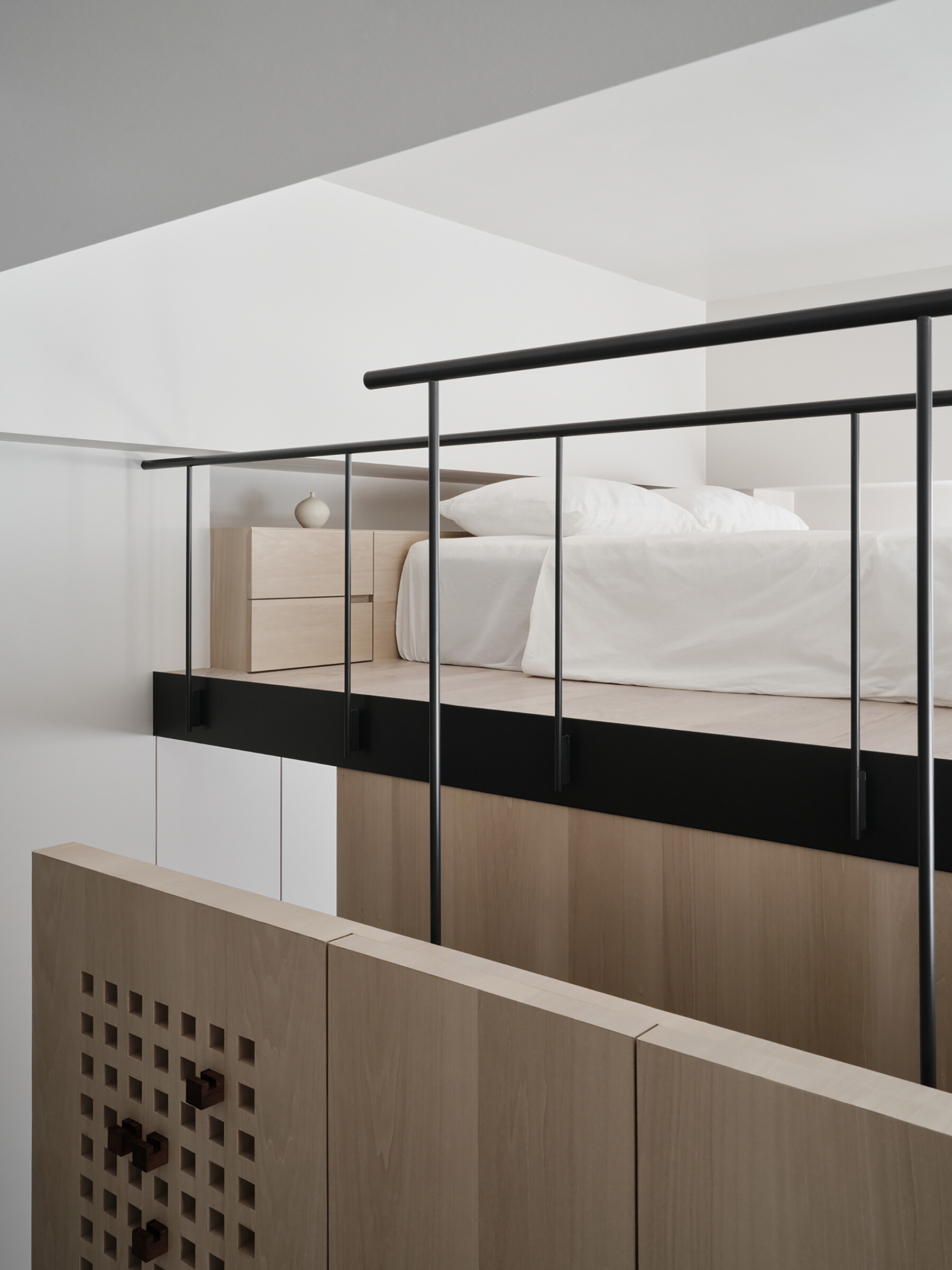 Soft, pale wood tones and clean lines define this clever loft setup, where every inch is thoughtfully designed. The built-in structure combines a TV wall, concealed storage, and a raised sleeping area above, all bathed in gentle natural light. Matte black railings and track lighting add a sharp contrast, giving the space a quietly modern edge. It’s a smart solution that feels both minimal and cozy, perfect for small-space living without ever feeling cramped.