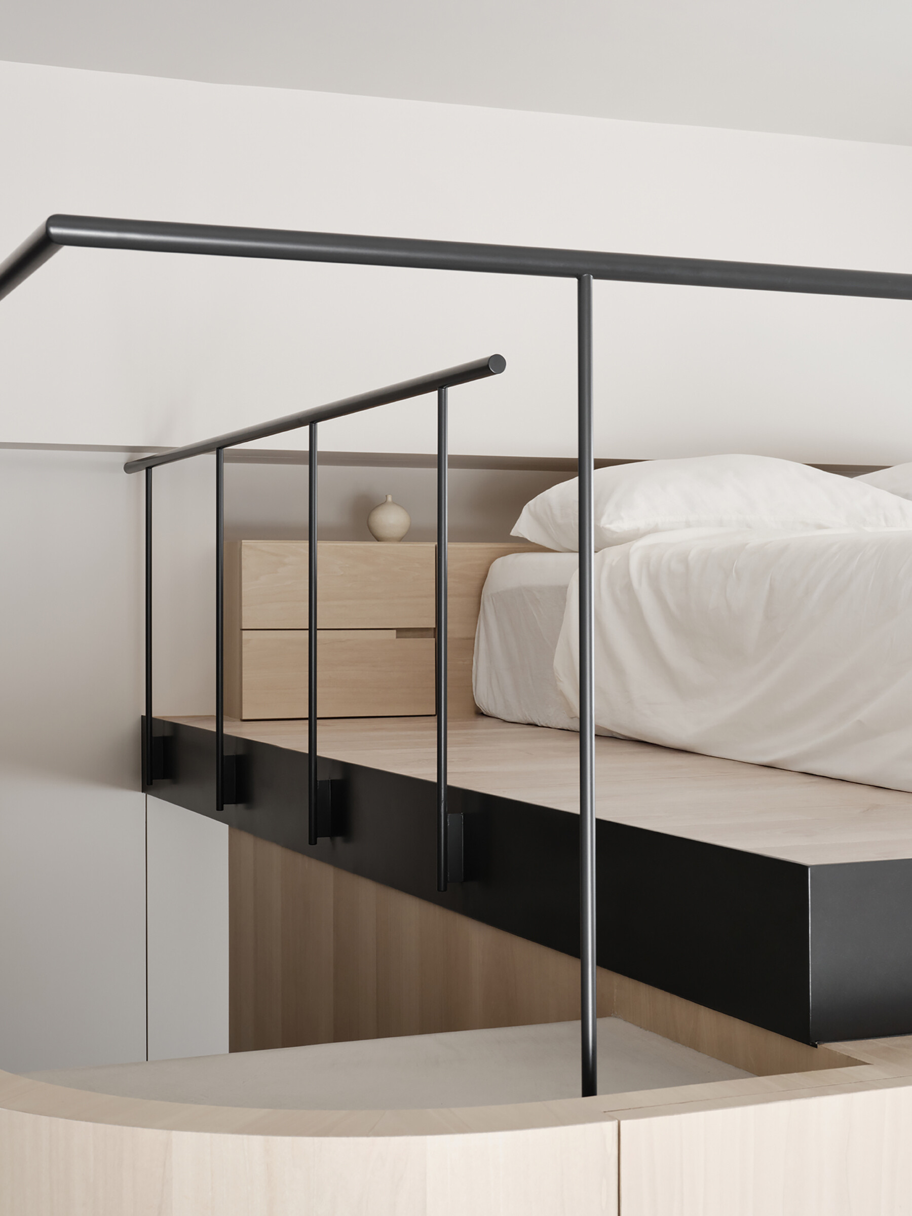 Soft, pale wood tones and clean lines define this clever loft setup, where every inch is thoughtfully designed. The built-in structure combines a TV wall, concealed storage, and a raised sleeping area above, all bathed in gentle natural light. Matte black railings and track lighting add a sharp contrast, giving the space a quietly modern edge. It’s a smart solution that feels both minimal and cozy, perfect for small-space living without ever feeling cramped.