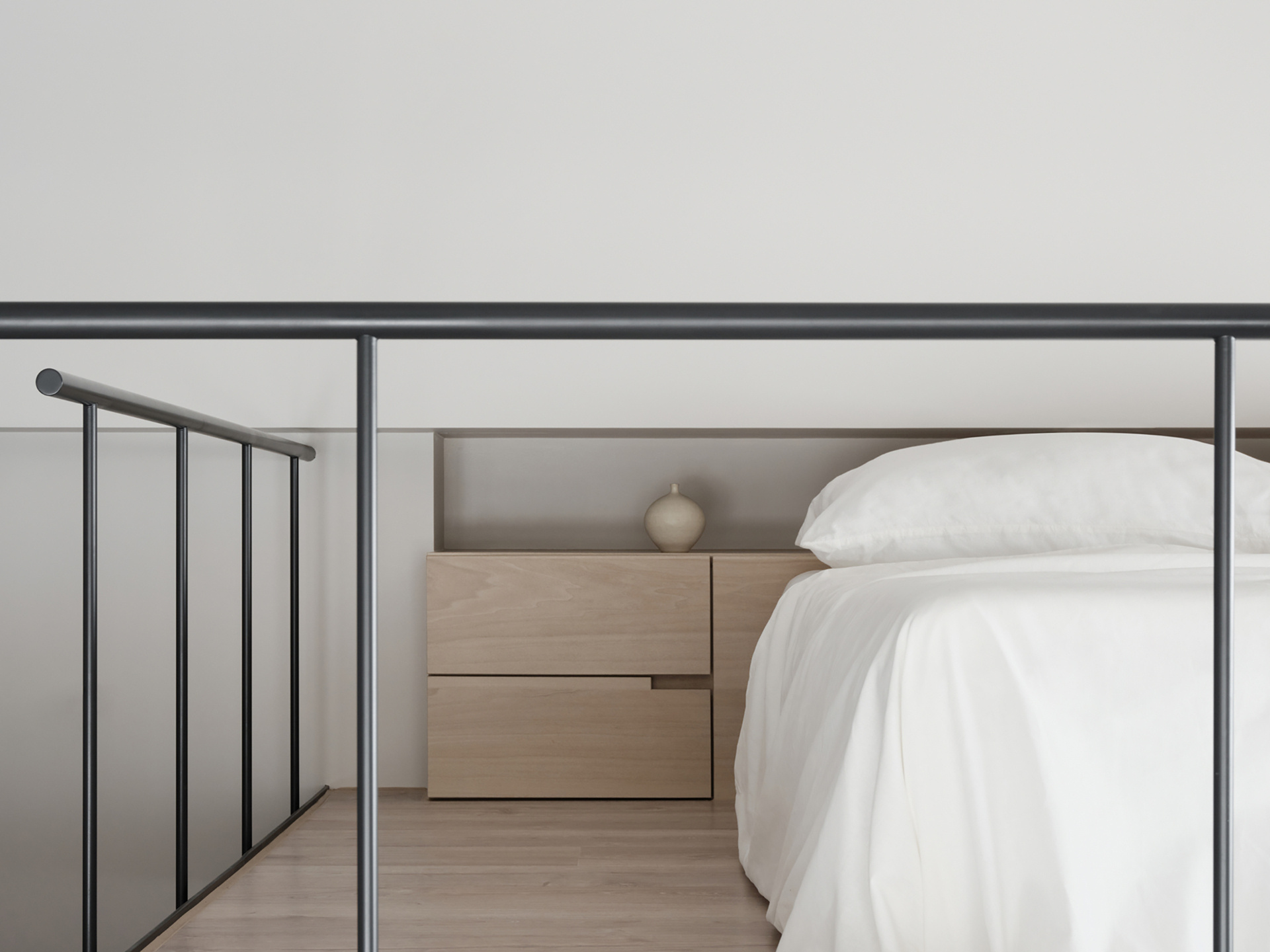 Soft, pale wood tones and clean lines define this clever loft setup, where every inch is thoughtfully designed. The built-in structure combines a TV wall, concealed storage, and a raised sleeping area above, all bathed in gentle natural light. Matte black railings and track lighting add a sharp contrast, giving the space a quietly modern edge. It’s a smart solution that feels both minimal and cozy, perfect for small-space living without ever feeling cramped.