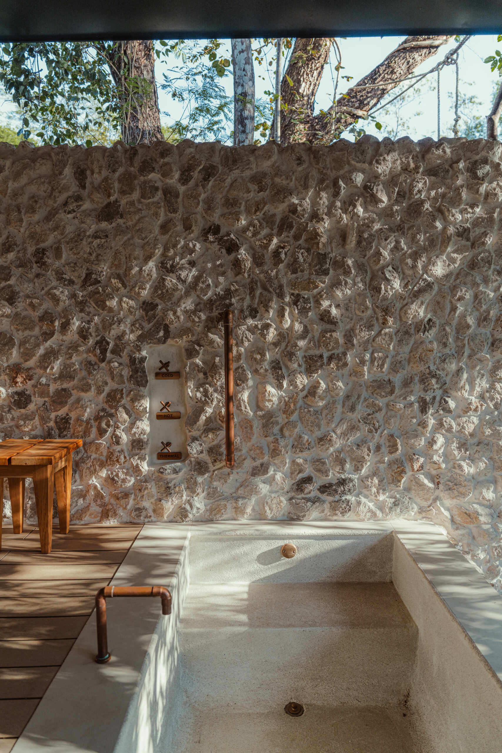 This outdoor bath feels like a hidden retreat carved into nature itself. Rough-cut stone walls catch the shifting sunlight, casting textured shadows that dance across the scene. The deep soaking tub, with its sand-toned finish and exposed copper fittings, brings a raw simplicity that’s quietly elegant.
