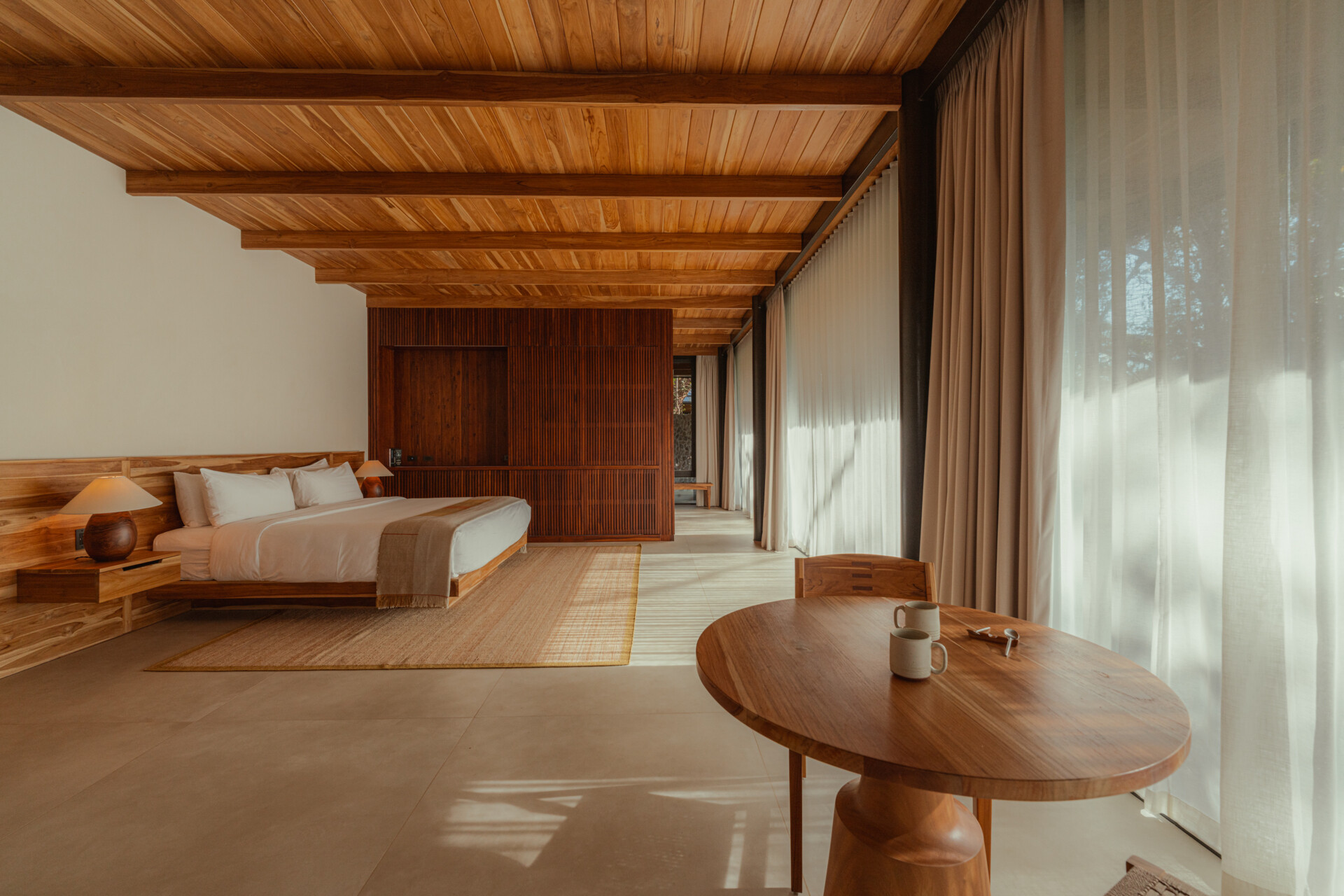 This warm, light-filled guest accommodation invites rest and relaxation. Sheer curtains, natural wood paneling and ceiling, and sculpted side tables create a calm, handcrafted feel, while the low bed and open layout make space for slow, restful moments.