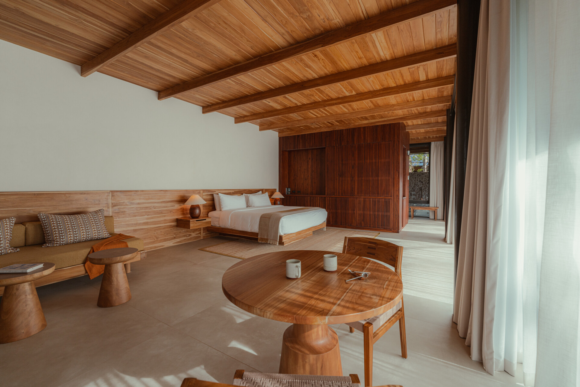 This warm, light-filled guest accommodation invites rest and relaxation. Sheer curtains, natural wood paneling and ceiling, and sculpted side tables create a calm, handcrafted feel, while the low bed and open layout make space for slow, restful moments.