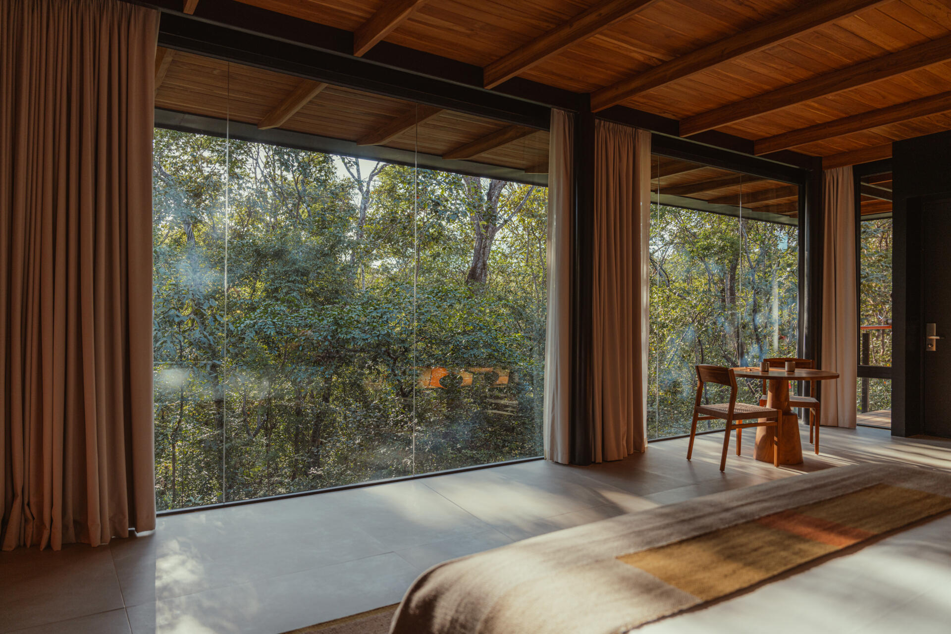 Golden light fills this accommodation, where a round wood table and woven chairs offer a quiet spot to pause. Surrounded by glass and soft curtains, the treetop view and natural textures create a grounded, peaceful atmosphere.