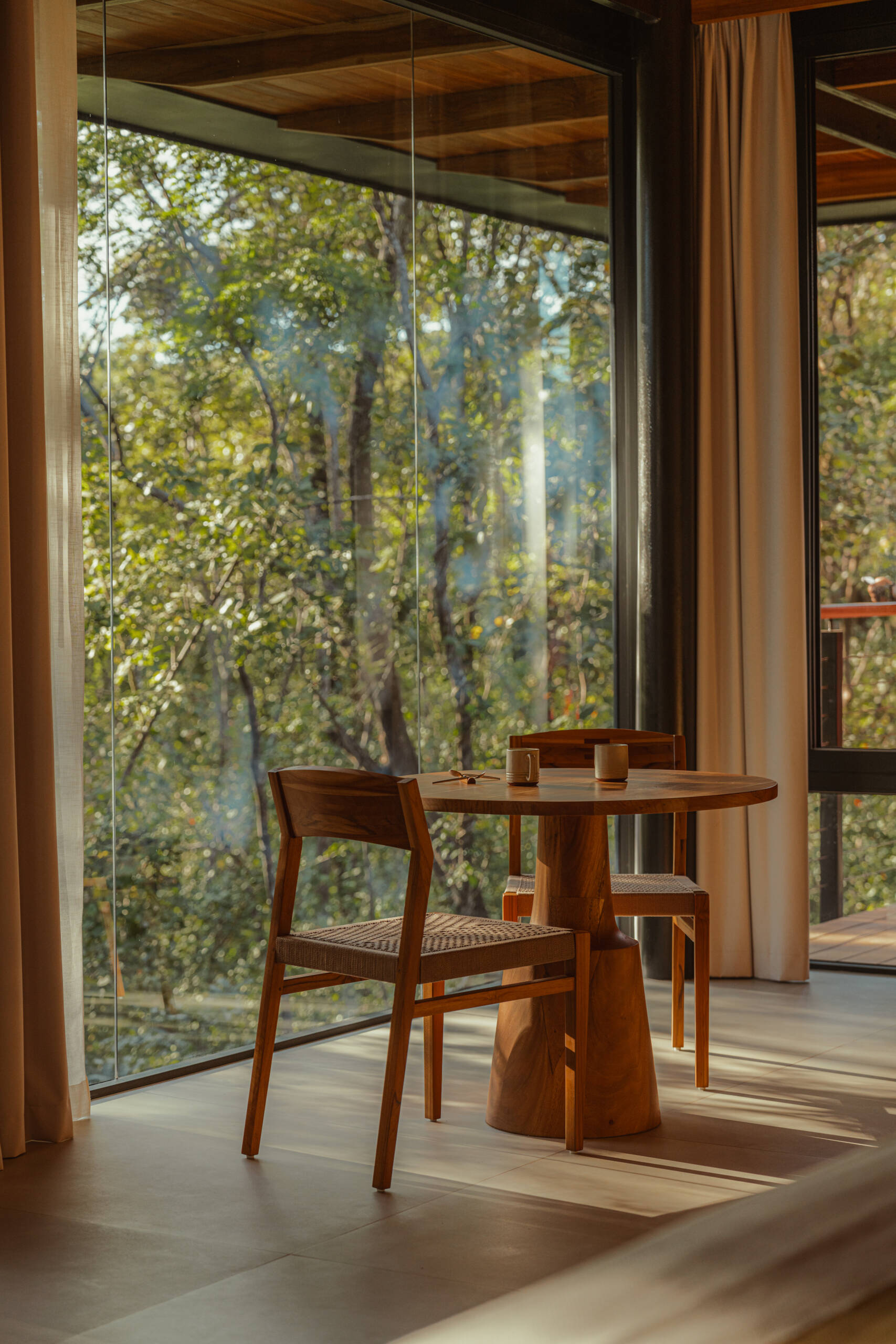 Golden light fills this small dining area, where a round wood table and woven chairs offer a quiet spot to pause. Surrounded by glass and soft curtains, the treetop view and natural textures create a grounded, peaceful atmosphere.
