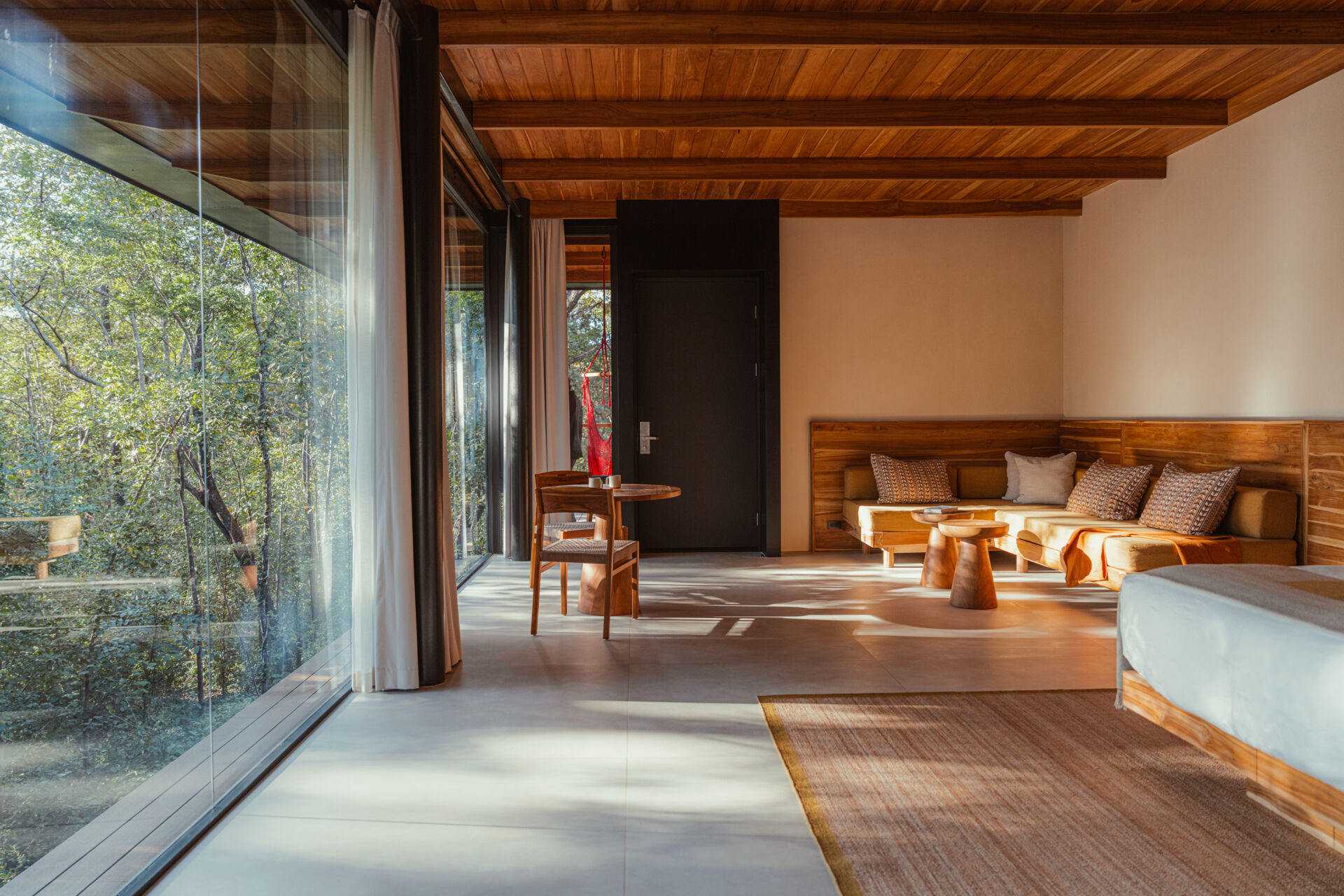 Sunlight pours through floor-to-ceiling glass, warming the pale tile floor and lounge area. Forest views and earthy textures create a grounded, peaceful vibe, while soft pillows and a round table invite quiet conversation and slow, cozy moments.