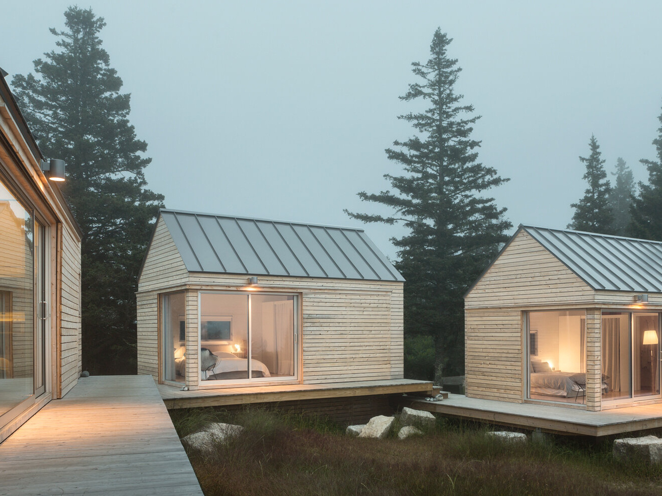 Set on a rocky outcrop and surrounded by evergreens, these three wood-clad cabins feel modern yet rooted in the landscape. Each simple, gabled form is wrapped in natural wood and topped with a metal roof, blending seamlessly with the forest and stone.

Sliding glass doors open to wide views and soft light, while a low wooden platform links the cabins as both walkway and gathering space. The design is quiet and deliberate, letting the rugged setting speak while offering a calm place to stay.