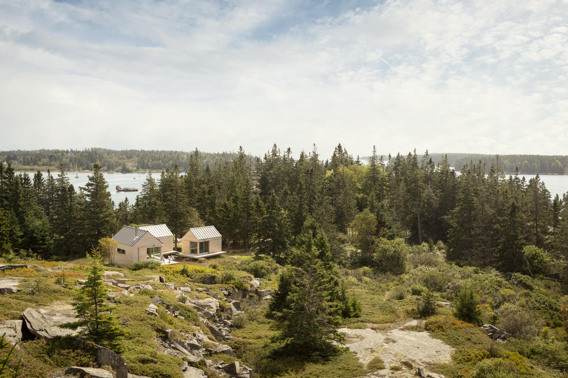 Set on a rocky outcrop and surrounded by evergreens, these three wood-clad cabins feel modern yet rooted in the landscape. Each simple, gabled form is wrapped in natural wood and topped with a metal roof, blending seamlessly with the forest and stone.
Sliding glass doors open to wide views and soft light, while a low wooden platform links the cabins as both walkway and gathering space. The design is quiet and deliberate, letting the rugged setting speak while offering a calm place to stay.